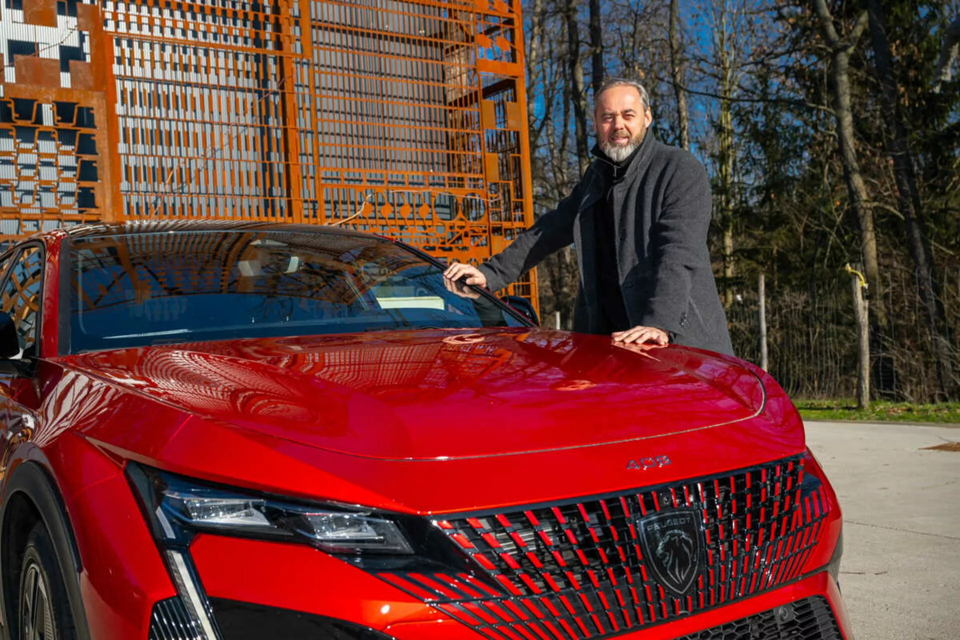 Modelo vermelho do Peugeot 408 de 2023 exibindo seu quarto frontal esquerdo à luz do dia, destacando os faróis LED detalhados, grade geométrica e capô esportivo e aerodinâmico com um homem de casaco escuro ao lado colocando sua mão no capô