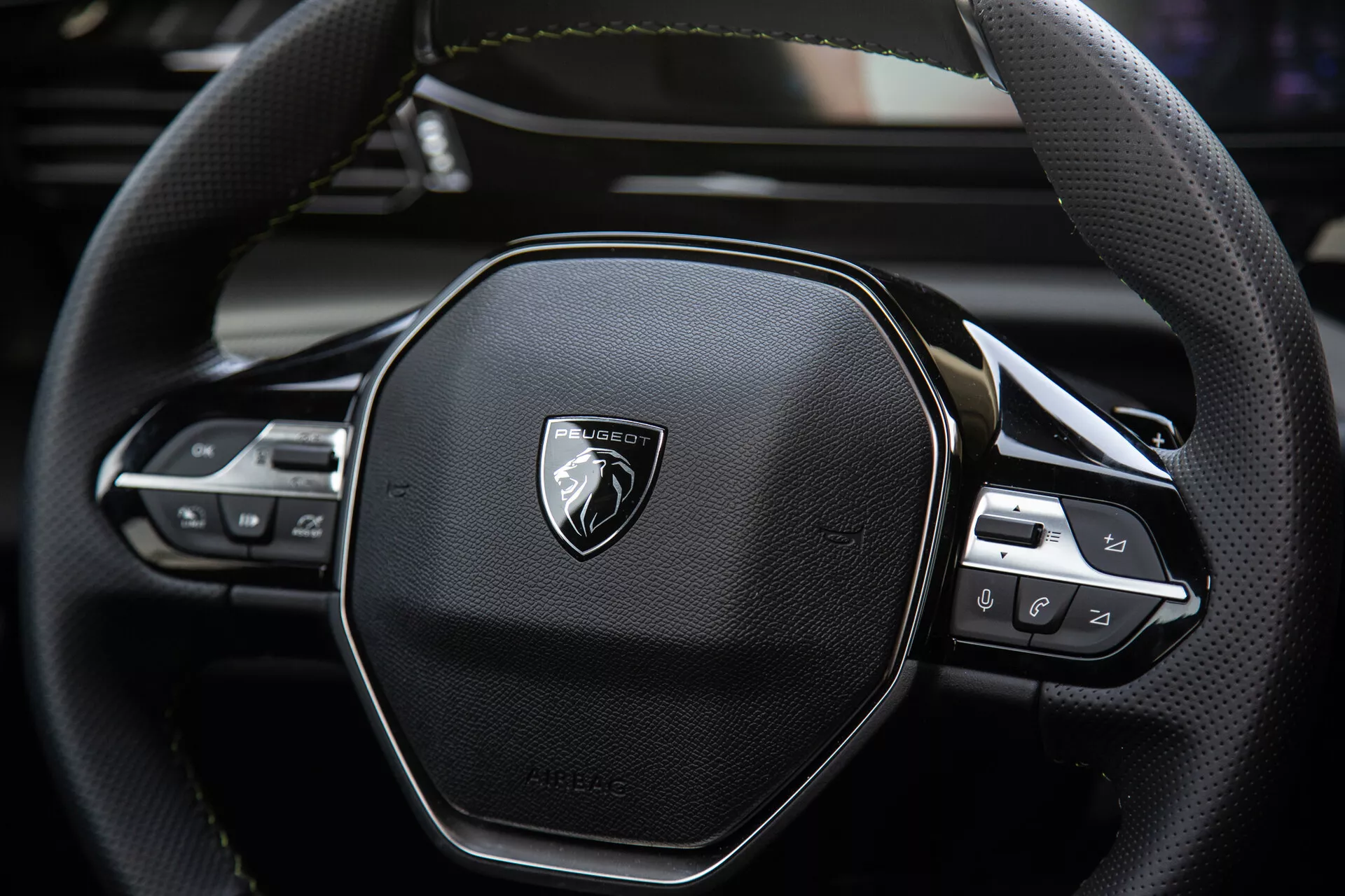Vista de perto do volante do Peugeot 408 2023 mostrando design ergonômico hexagonal, costura de couro, controles de tecnologia integrados para áudio e configurações de cruzeiro, com emblema em metal escovado.