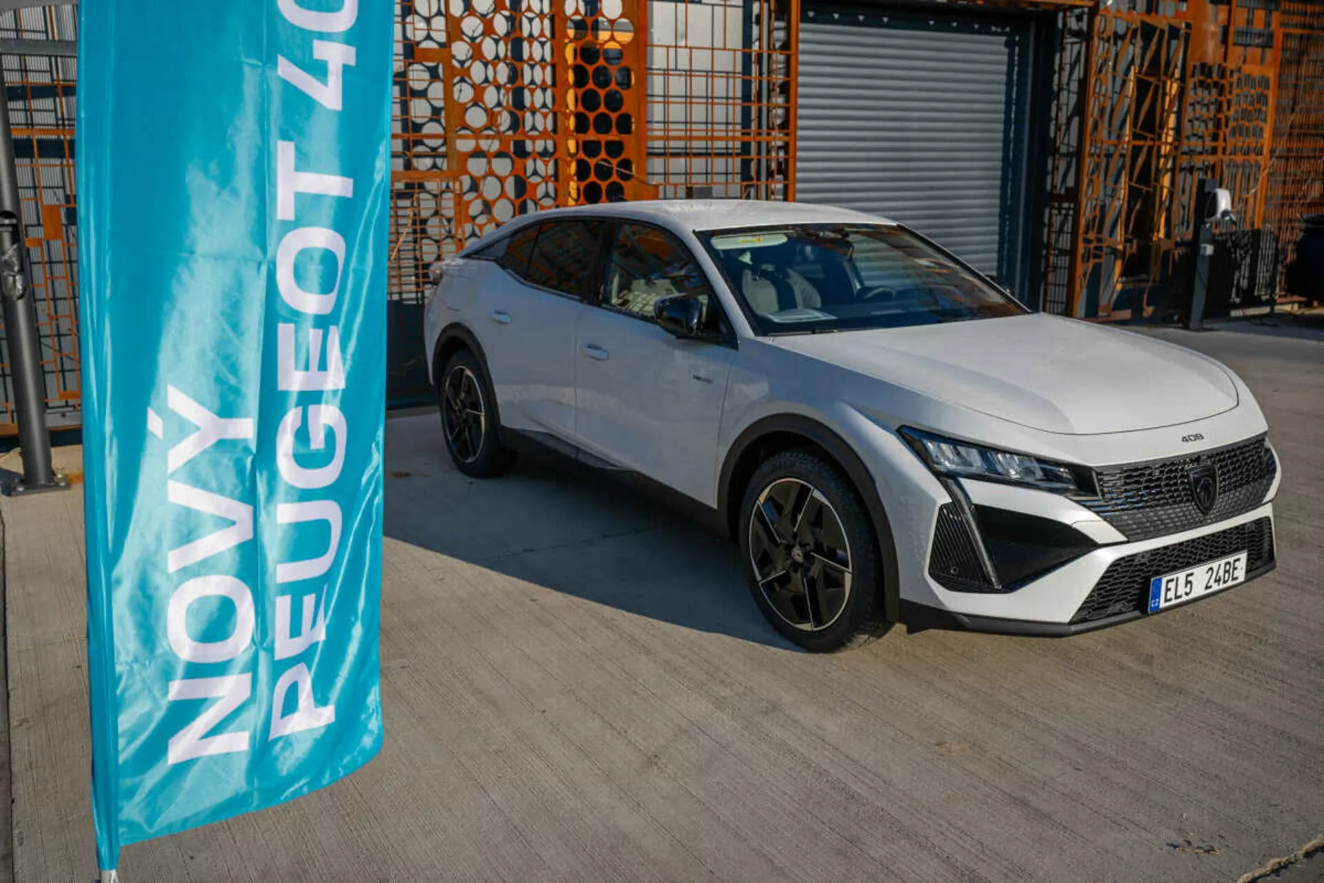 Peugeot 408 branco de 2023 estacionado ao ar livre com arquitetura industrial ao fundo, exibindo os recursos de design e tecnologia do veículo sob a luz natural do dia