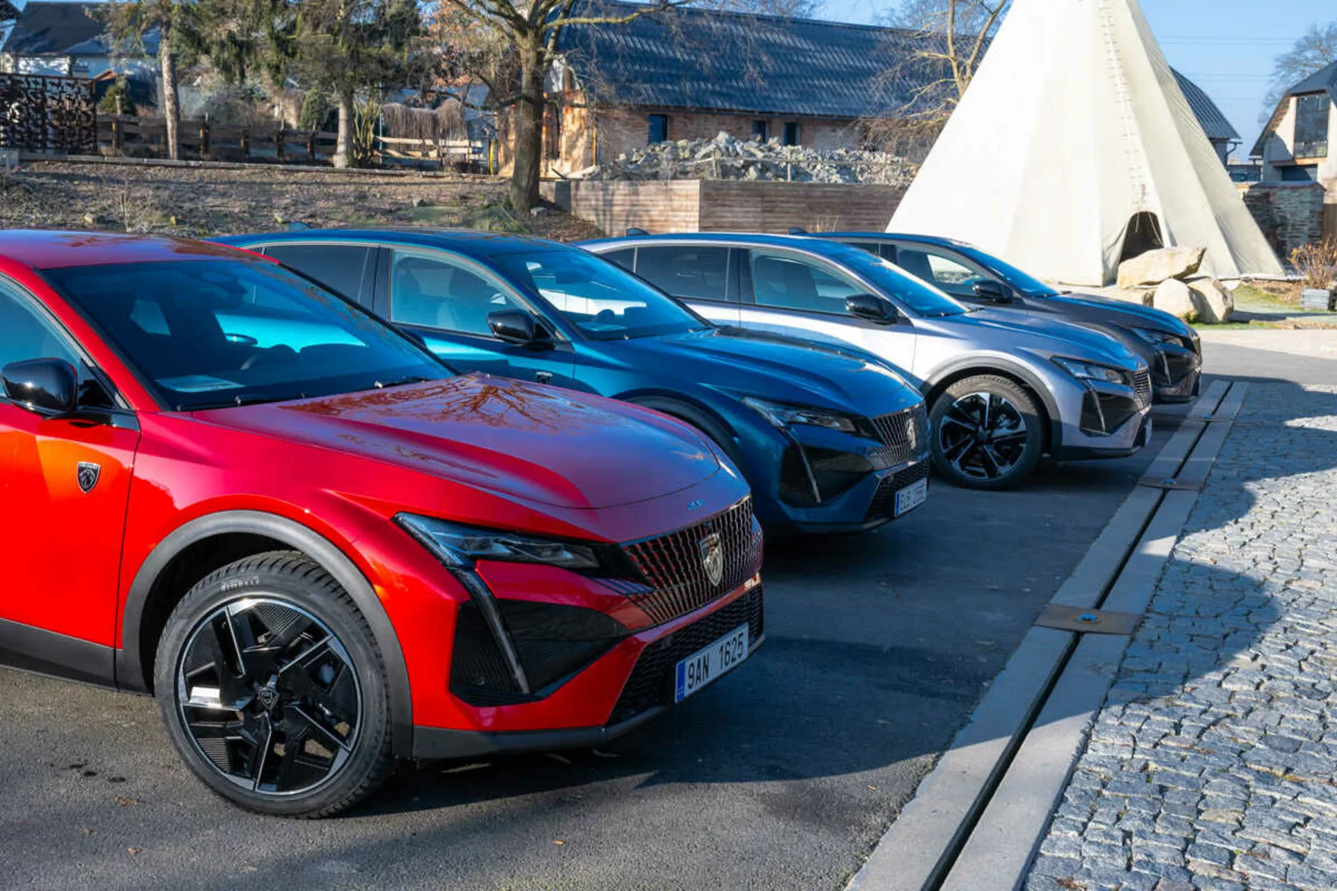 Linha de Peugeot 408 de 2023 com carro vermelho em primeiro plano com rodas esportivas multi-raios e faróis LED indicativos de modelos elétricos ou híbridos, estacionado em área pavimentada ao lado de calçada