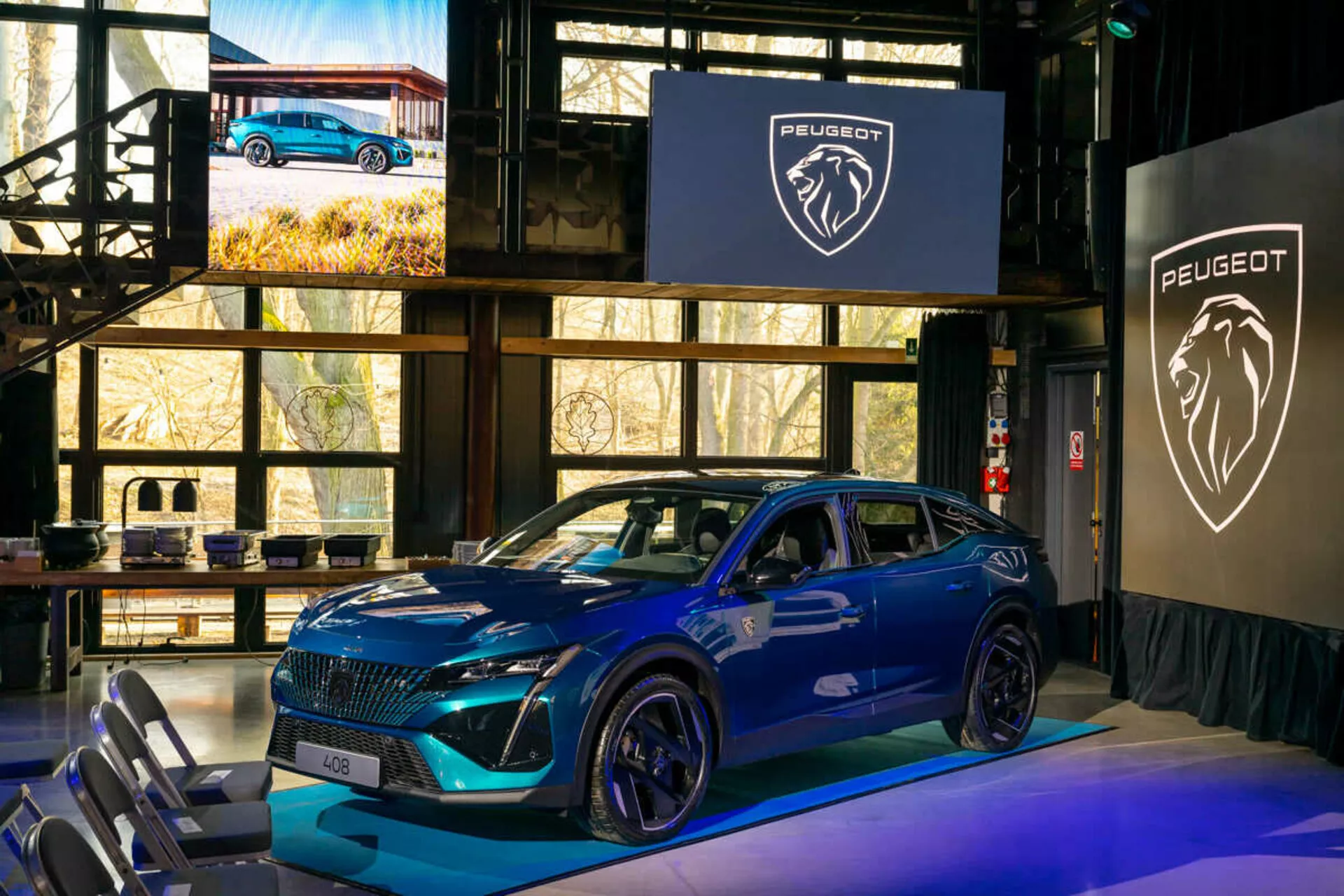 Peugeot 408 azul de 2023 apresentado em um evento interno, com uma grade distintiva, faróis LED afiados, capô esportivo contornado com entradas de ar, grandes rodas de liga leve e um perfil lateral dinâmico com indicadores de direção integrados nos espelhos laterais montados na porta