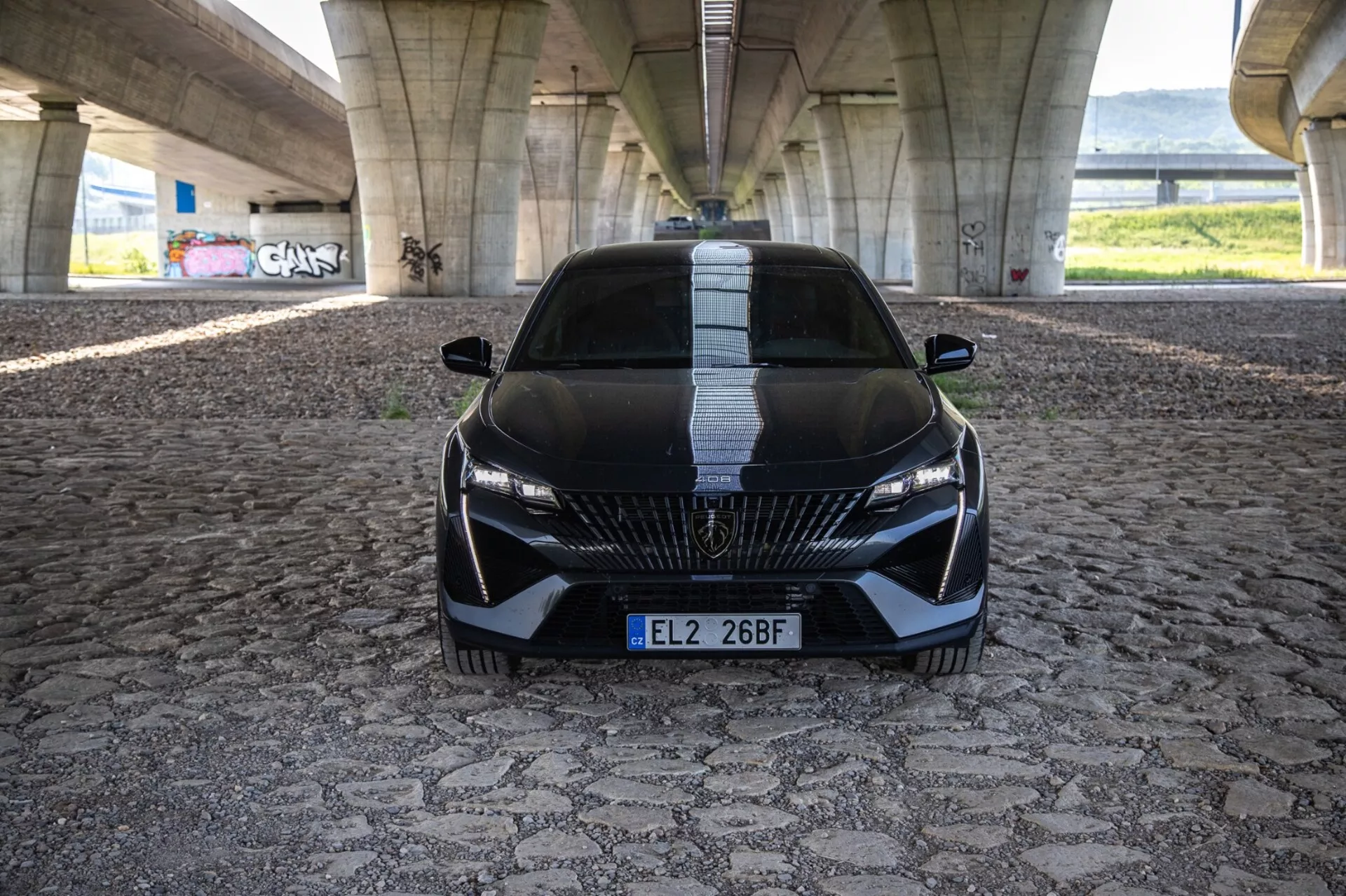 Peugeot 408 de 2023 em uma vista frontal dinâmica mostrando sua postura agressiva e design esportivo com grade detalhada, faróis LED elegantes e recursos aerodinâmicos em um pavimento padronizado sob uma ponte urbana