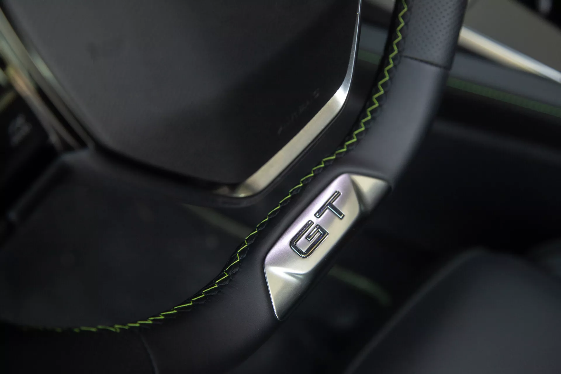 Vista detalhada da seção do volante e do painel do Peugeot 408 2023, apresentando um volante de couro preto com costura verde e um emblema GT, evidenciando luxo e esportividade