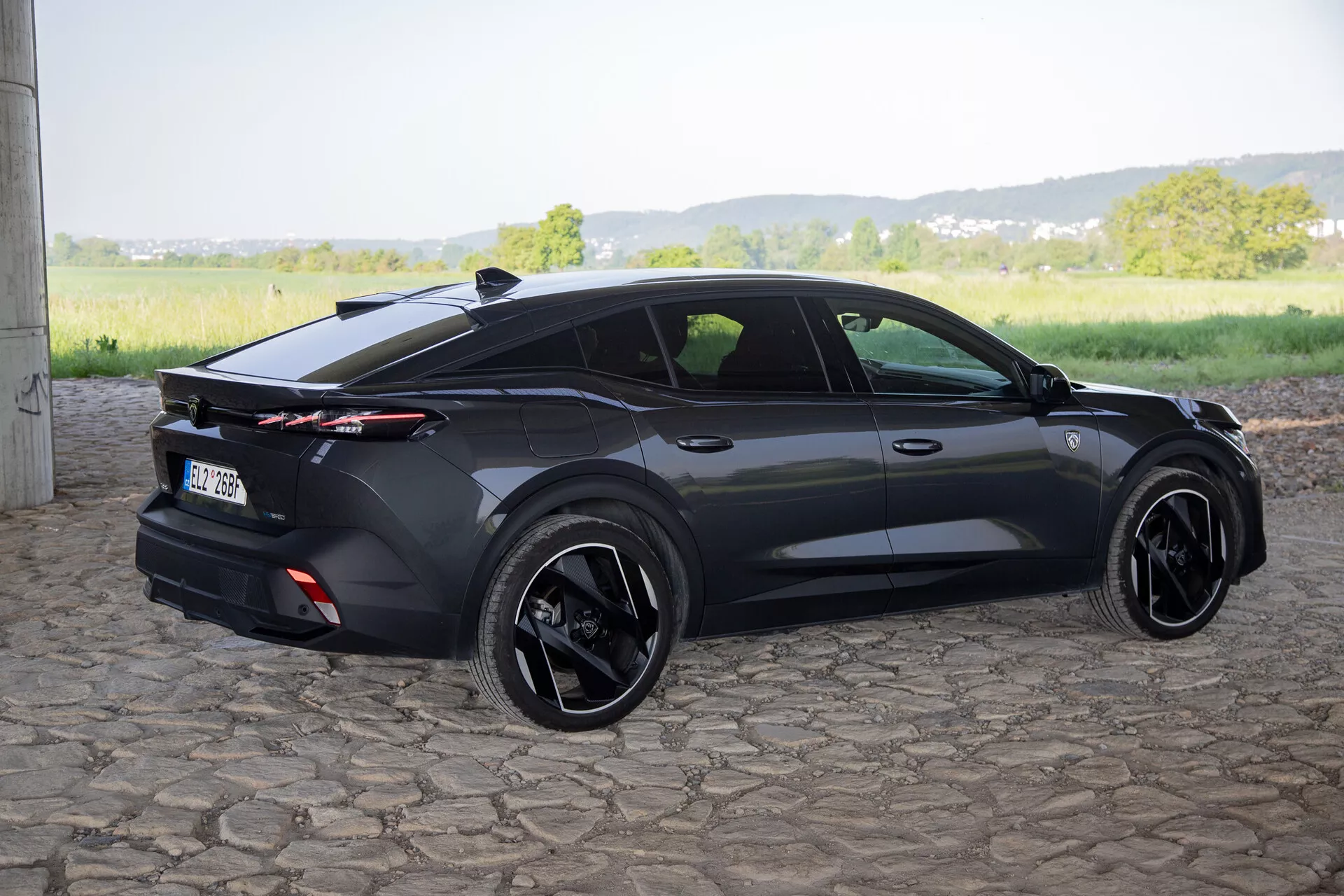 Peugeot 408 de 2023 captado de um ângulo traseiro de três quartos destacando seu design esportivo hatchback, spoiler traseiro integrado com luzes traseiras de comprimento total, rodas de liga leve pretas e características contemporâneas como maçanetas embutidas e espelhos laterais com setas, à luz do dia natural