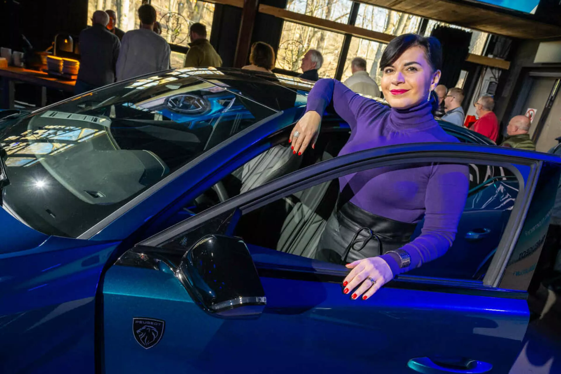 Carro esportivo azul vibrante, possivelmente um Lamborghini, capturado de um ângulo de três quartos enfatizando suas linhas elegantes, faróis LED afiados e icônico logotipo em forma de escudo, com uma pessoa interagindo casualmente com o veículo sugerindo luxo e estilo.