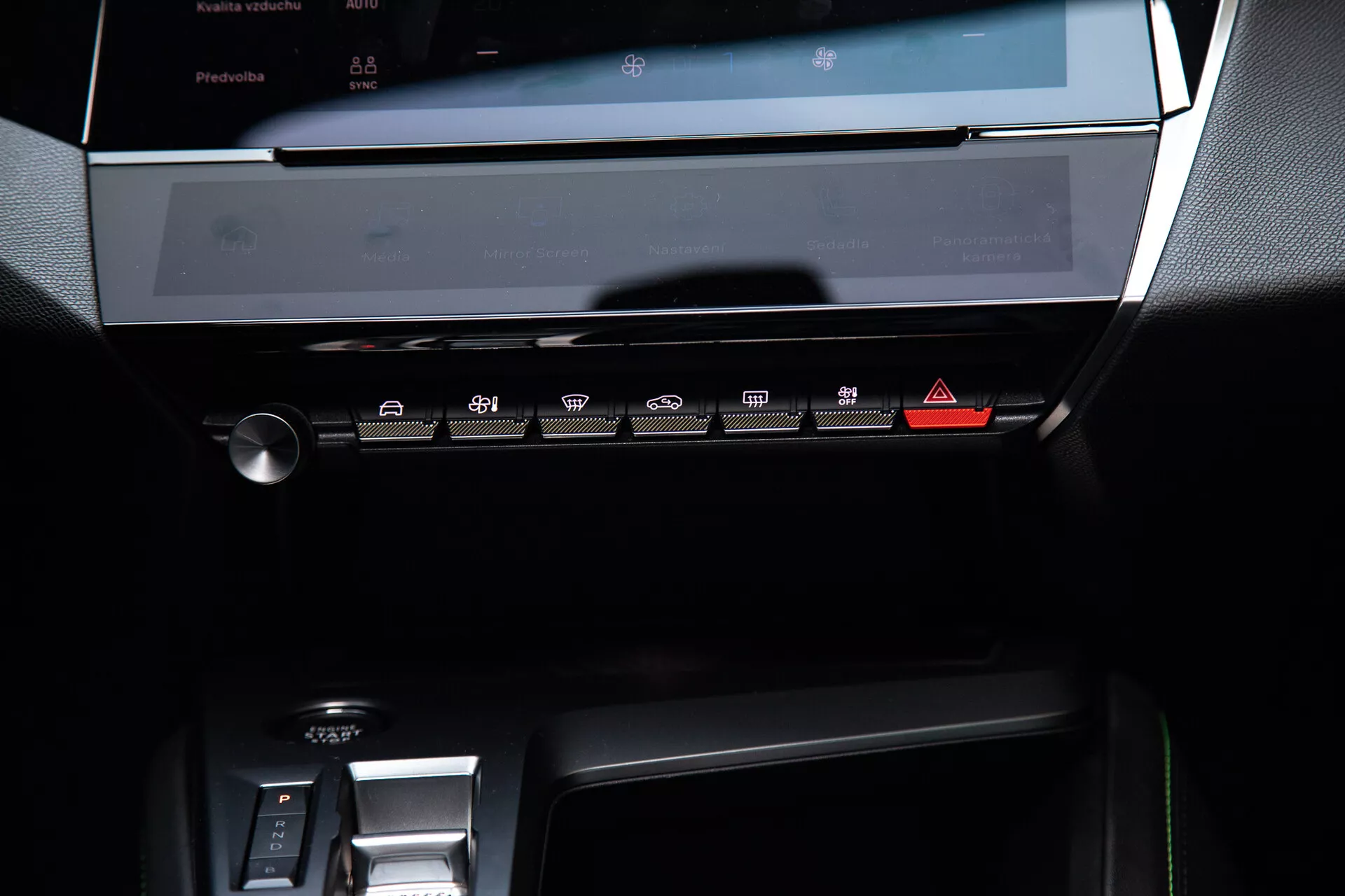 Painel central do Peugeot 408 2023 exibindo o sistema de infoentretenimento com tela touchscreen desligada, painel de controle climático com botões sensíveis ao toque e parte da área de mudança de marchas eletrônica, sob uma suave iluminação ambiente destacando seu design moderno e elegante com tons escuros e acabamentos metálicos.