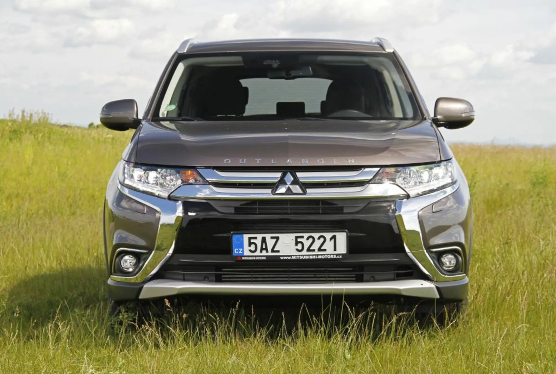 Imagem de frente de um Mitsubishi Outlander posicionado em um campo gramado destacando sua grade frontal Dynamic Shield, faróis LED e design robusto do para-choque, mostrando capacidade off-road e características tecnológicas modernas durante o dia.
