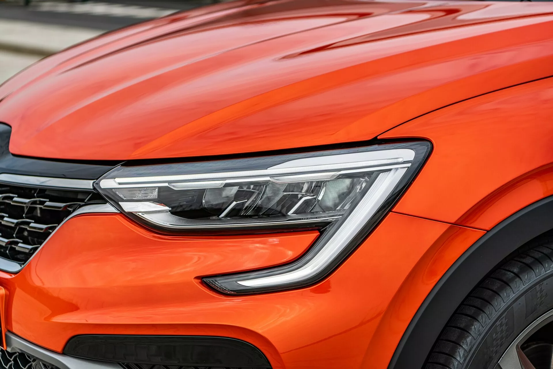 Vista aproximada da parte frontal de um brilhante Renault Arkana 2021 laranja, focada nos detalhes de design modernos incluindo faróis LED avançados, curvas aerodinâmicas do capô, uma grelha escura parcialmente visível com padrão em favo de mel, e um fragmento de uma grande roda de liga, sugerindo desempenho premium e tecnologia automotiva de ponta.
