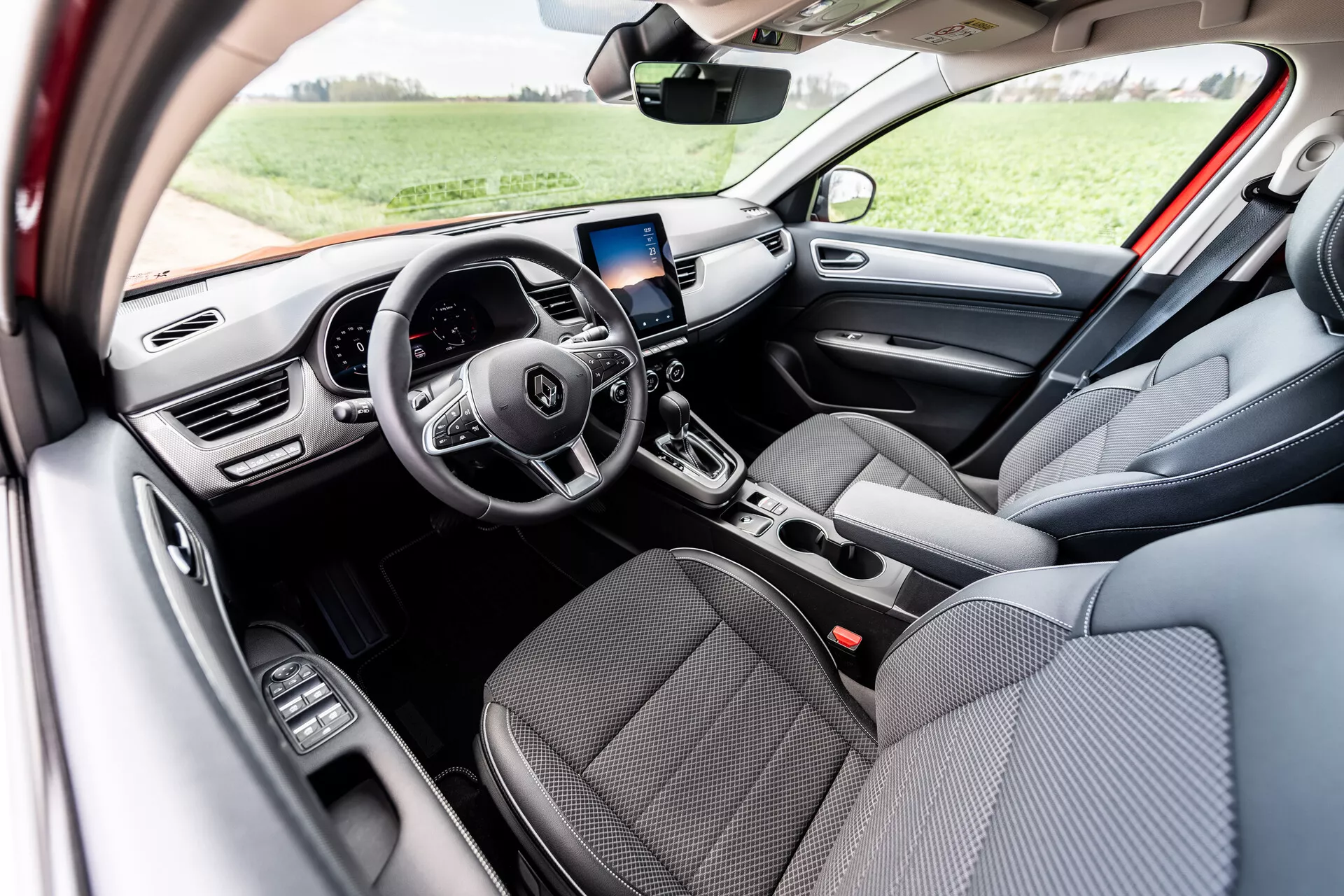 Vista interior do Renault Arkana 2021 exibindo assentos dianteiros em tecido cinza, volante multifuncional de três raios com botões de controle integrados, display digital no painel, tela de infotainment com navegação GPS acima das saídas de ar centrais, controles manuais de ar-condicionado, alavanca de câmbio automático e controles de vidros elétricos na porta do lado do motorista, iluminada pela luz natural do dia em um ambiente externo