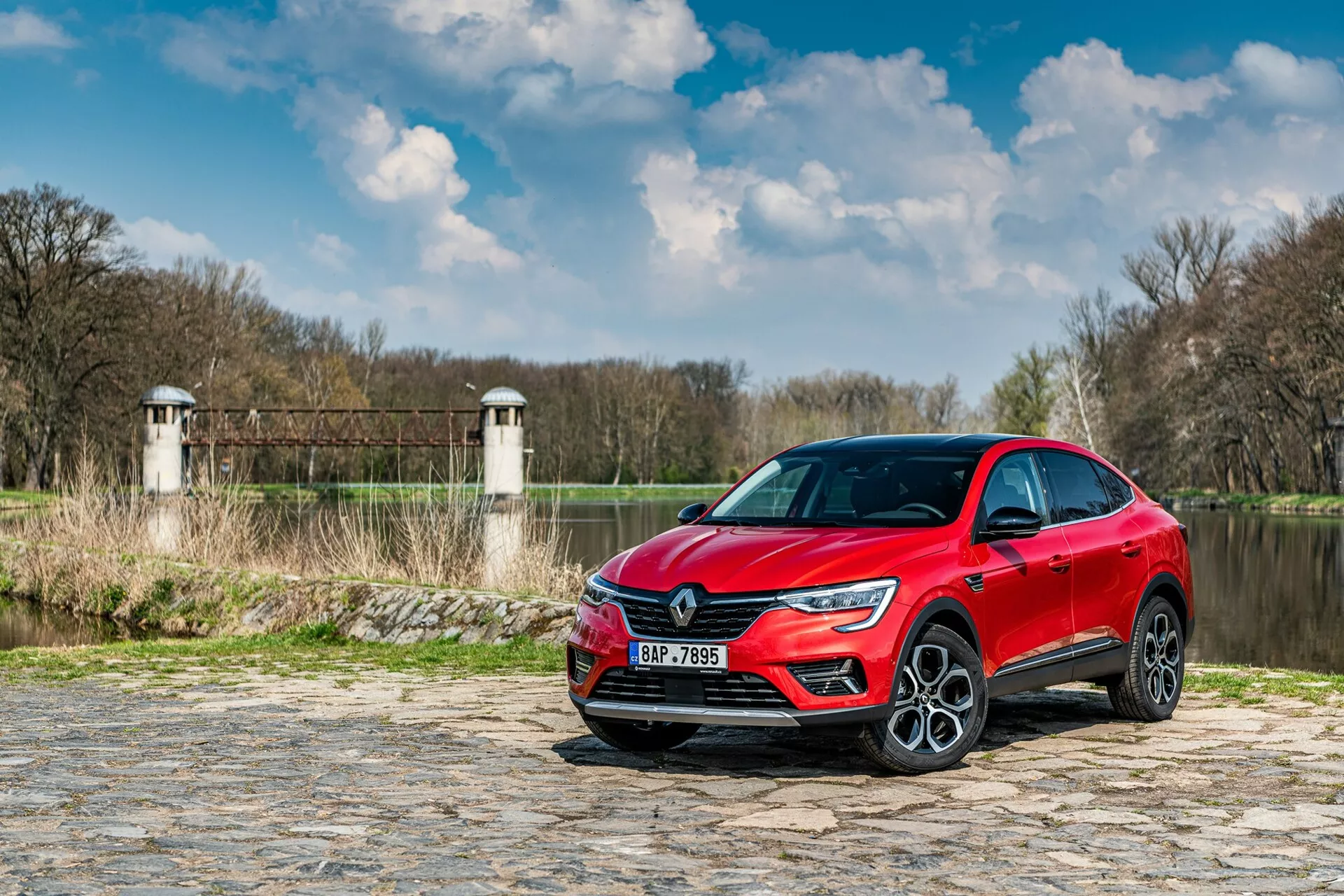 A imagem retrata um Renault Arkana 2021 estacionado em um caminho de paralelepípedos perto de um rio ou lago, com vegetação e estruturas que lembram torres de uma barragem ou ponte ao fundo. O carro é capturado de um ângulo baixo, vista frontal de três quartos, destacando a grade dianteira, faróis LED e parte do perfil lateral, incluindo a porta do motorista e o para-lama dianteiro. Os elementos de design moderno, como a assinatura de luz nítida dos LEDs e as grandes rodas de liga, sugerem um nível de acabamento superior. A pintura metálica reflexiva adiciona apelo estético, enquanto os possíveis sensores de assistência ao motorista e a tecnologia embutida nos para-choques e espelhos laterais melhoram a funcionalidade.
