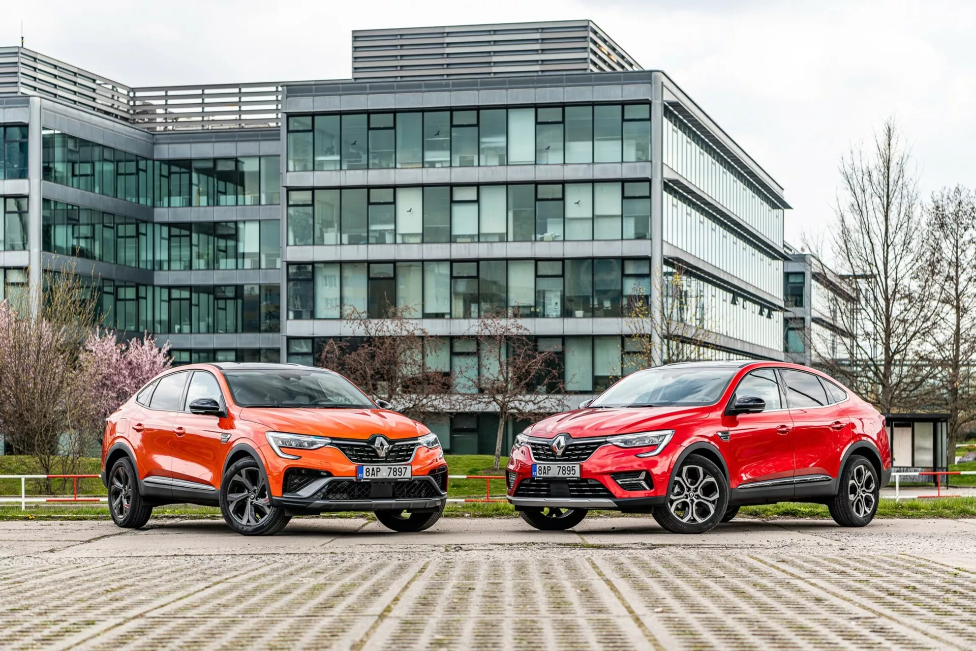 A imagem mostra dois veículos estilo SUV estacionados lado a lado em frente a um moderno edifício de escritórios com fachada de vidro. O veículo à esquerda está pintado em laranja vibrante com um brilho metálico e apresenta uma grade frontal distintiva, faróis LED nítidos, capô esculpido e grandes rodas de liga leve multirraiadas com pneus de perfil baixo. O carro à direita tem acabamento metálico vermelho, com uma silhueta geral semelhante, faróis nítidos, grade proeminente e elegantes rodas de liga grandes. Ambos os veículos exibem linhas angulares, espelhos aerodinâmicos e acabamentos inferiores contrastantes. A foto, tirada de um ângulo baixo próximo ao nível do solo, destaca o ambiente urbano e realça o design, a presença e as características tecnológicas avançadas dos veículos.