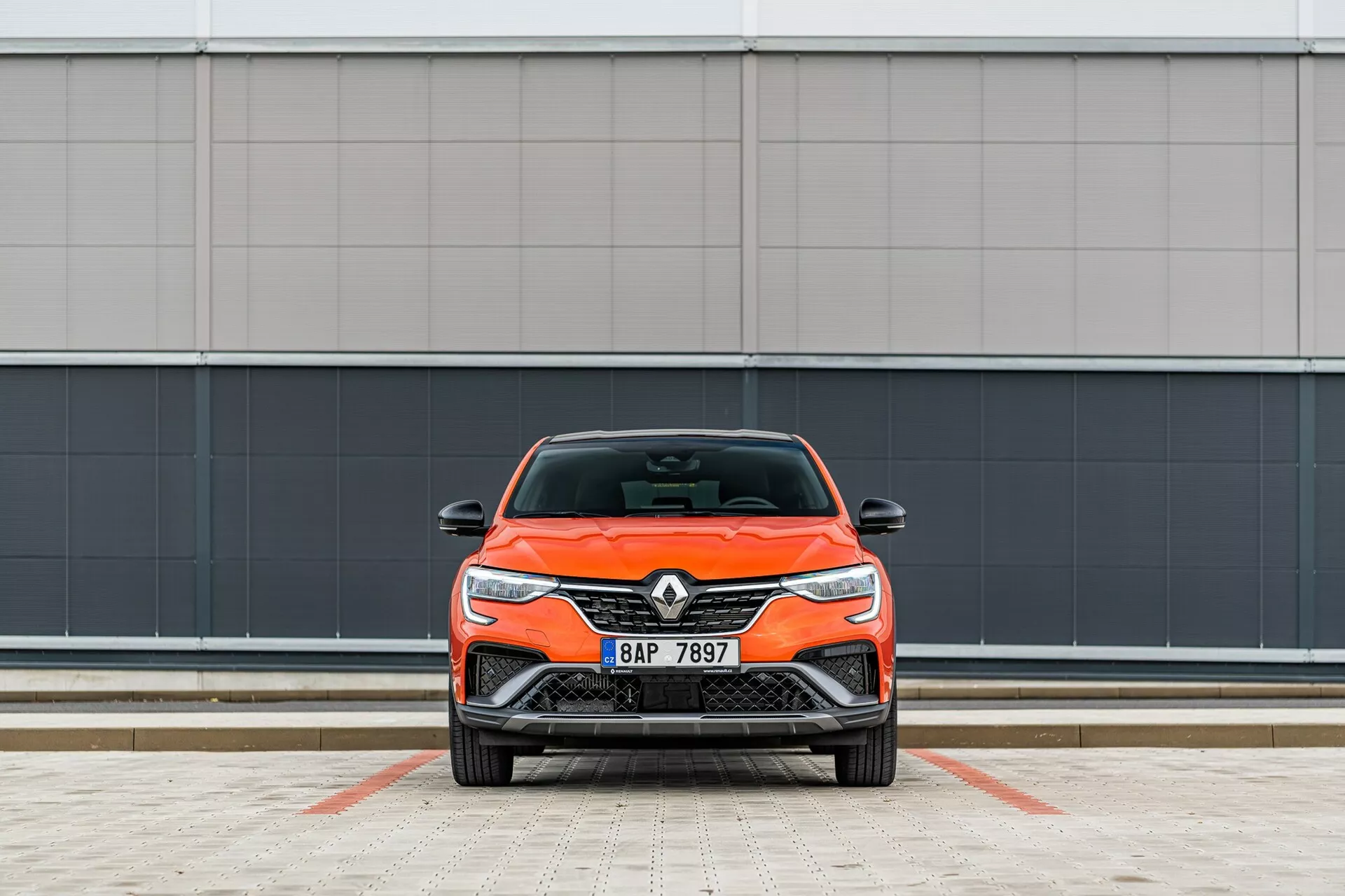 Vista frontal de um carro Renault laranja brilhante, destacando seu design moderno com faróis elegantes e grade frontal, estacionado perpendicularmente à câmera contra um fundo de parede cinza minimalista em uma área de estacionamento demarcada.