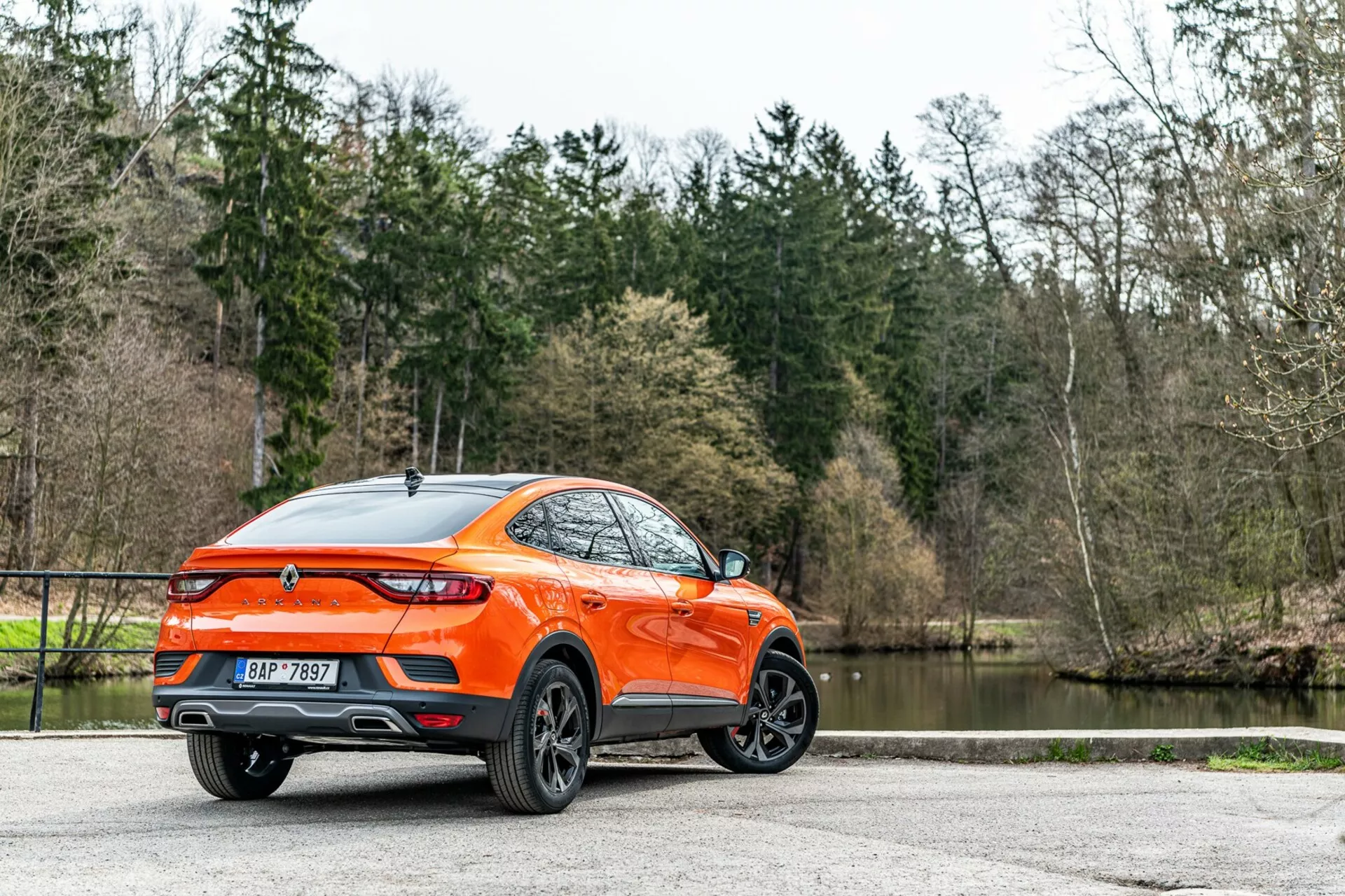 O Renault Arkana 2021 representado de um ângulo traseiro de três quartos, visão detalhada dos perfis lateral e traseiro, mostrando um crossover/SUV moderno com maior altura do solo e linha de teto esportiva tipo coupé. A carroceria laranja viva se destaca contra as árvores e o lago, destacando as rodas de liga leve de design multi-raios, luzes traseiras LED, spoiler traseiro, antena de barbatana de tubarão, emblema centralmente colocado, ponteiras de escape duplas, acabamento de tinta brilhante e vidros traseiros escurecidos. A iluminação natural difusa melhora a visibilidade do exterior bem-cuidado, sugerindo um apelo para consumidores com estilos de vida ativos, estacionado em uma superfície pavimentada.