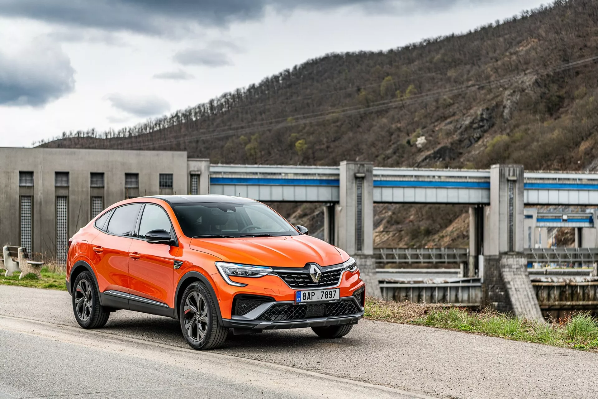 Um moderno crossover SUV Renault Arkana 2021 de cor laranja brilhante está estacionado ao lado de uma estrada, fotografado de um ângulo lateral frontal focando no lado do motorista. A imagem mostra a grade frontal do carro com um grande emblema, faróis angulares e elegantes de LED, luzes diurnas, entradas de ar e possivelmente sensores de assistência ao motorista no para-choque inferior, arcos das rodas pronunciados com rodas de liga leve de múltiplos raios e espelhos com luzes de indicação de direção integradas. O veículo, classificado como SUV, tem uma postura elevada com maior distância ao solo e uma linha de teto inclinada em estilo cupê. Ao fundo, vê-se uma infraestrutura que se assemelha a uma represa ou sistema de comportas, situando o veículo num ambiente industrial ou rural. A foto é tirada de um ângulo ligeiramente baixo, destacando a altura e a postura do carro.