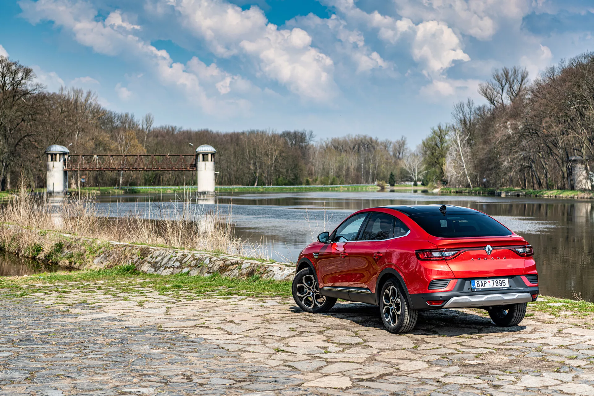 Renault Arkana 2021 estacionado em calçada perto da água com fundo natural, mostrando vista traseira de três quartos, design esportivo vermelho e luzes traseiras LED