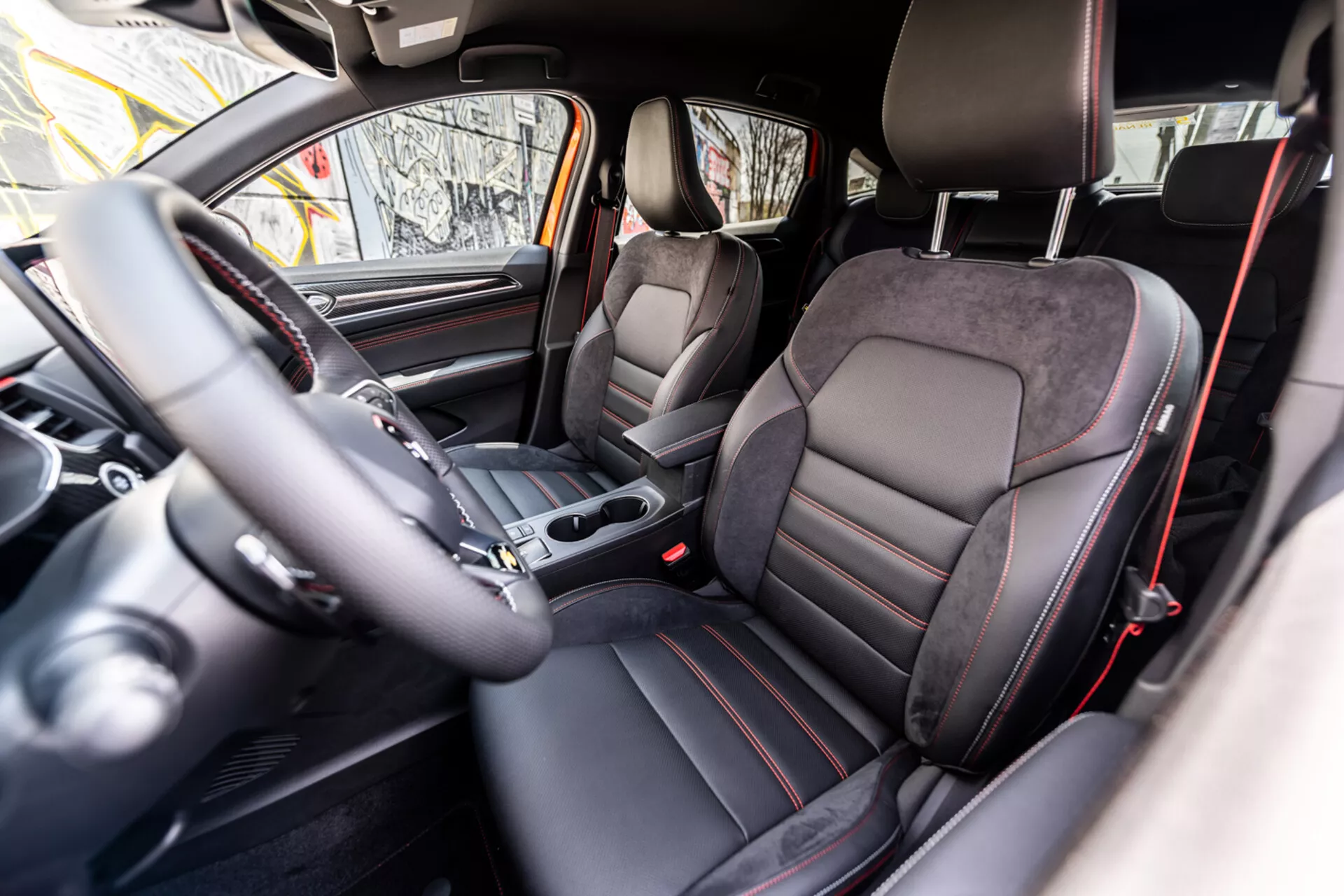 Interior do Renault Arkana 2021 mostrando a cabine frontal a partir da perspectiva do assento traseiro com assentos em estilo desportivo em couro e Alcantara, volante à esquerda com controles montados, alavanca de câmbio automático no console central, cluster de instrumentos analógico, tela integrada do sistema de infotenimento e esquema de cores escuras premium com detalhes metálicos