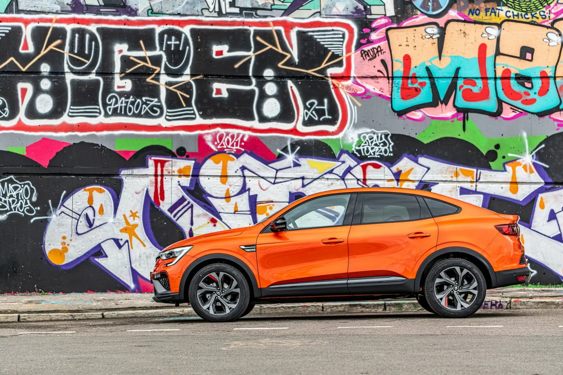 A imagem mostra um crossover SUV Renault Arkana 2021 de cor laranja vibrante estacionado paralelo a uma rua com uma parede de graffiti de cores vivas ao fundo. Capturada de um ângulo de visão lateral, a fotografia destaca o design esportivo e moderno do veículo com portas dianteiras e traseiras do lado do motorista visíveis, janelas laterais, espelhos laterais e linha de teto inclinada. As rodas de liga estilizadas com pneus de perfil baixo sugerem um foco no desempenho em estrada. As maçanetas com entrada sem chave nas portas da frente indicam um sistema de entrada e partida sem chave. A imagem foi tirada à luz do dia, enfatizando o contraste da carroceria laranja contra o graffiti e mostrando a carroceria bem mantida e nítida do carro em um ambiente urbano e juvenil.