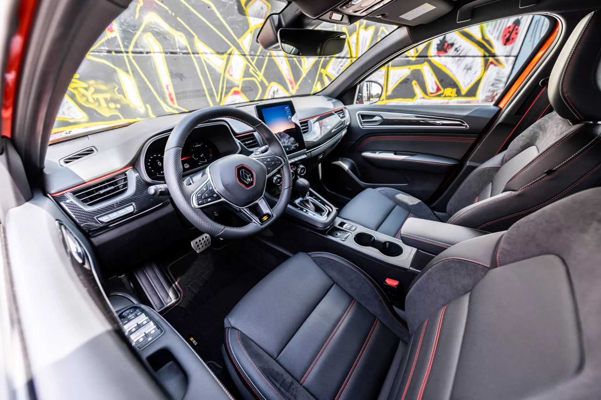 Interior do Renault Arkana 2021 mostrando a cabine dianteira luxuosa a partir da perspectiva da porta do passageiro com volante moderno de três raios, display sensível ao toque para o sistema de infotainment e estofamento esportivo com costuras vermelhas