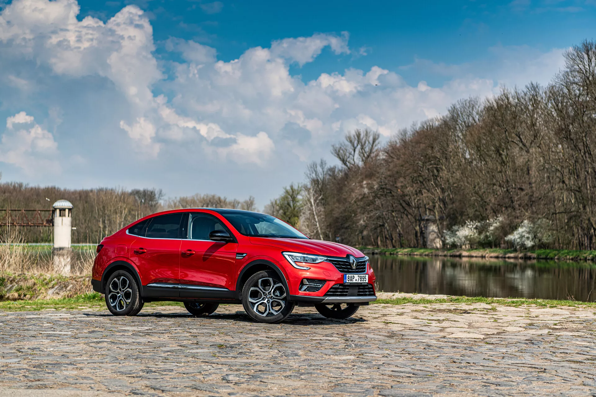 SUV vermelho Renault Arkana 2021 estacionado em pavimento de pedra com corpo de água e árvores ao fundo sob um céu parcialmente nublado, perfil lateral destaca seu design elegante e características dinâmicas