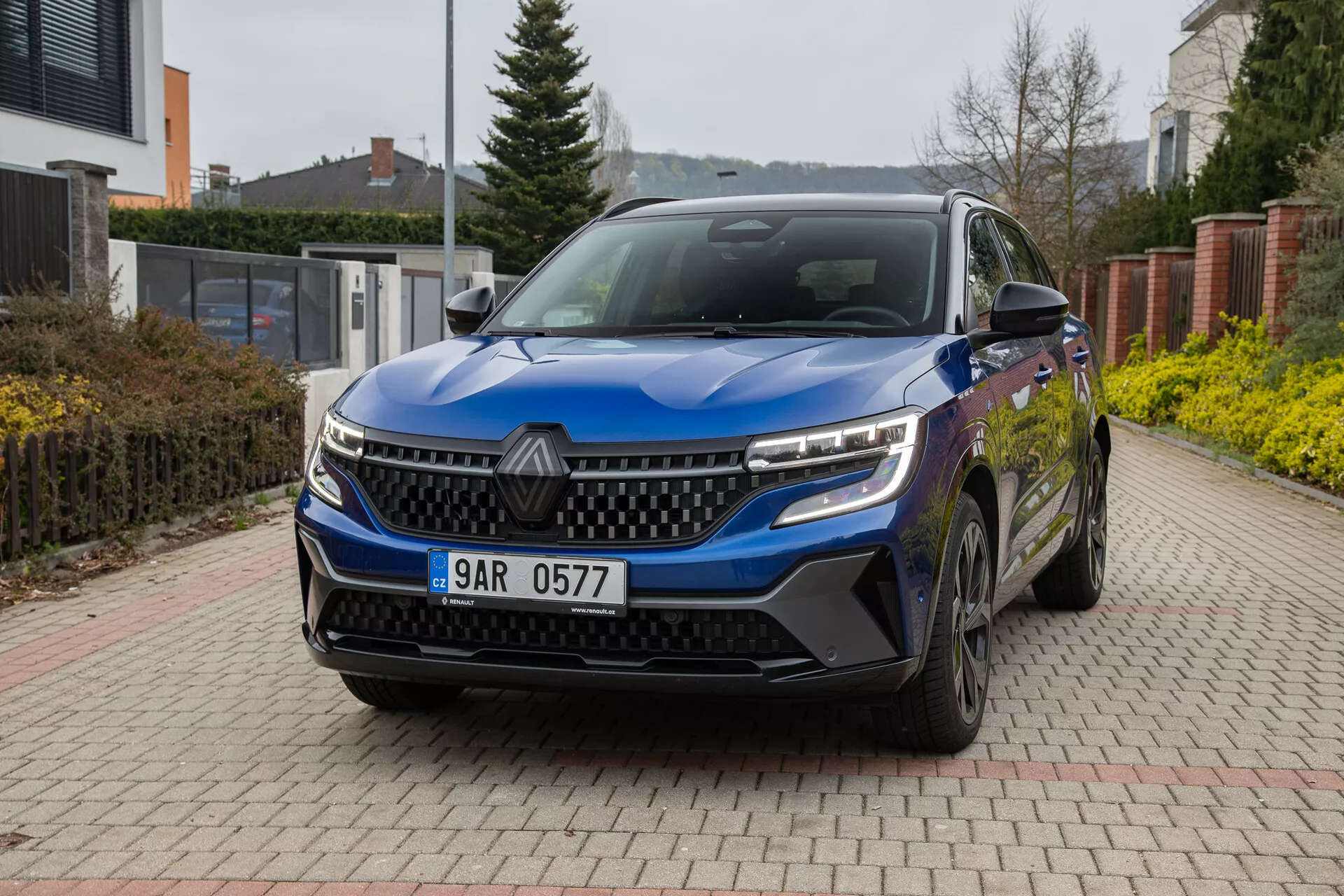 Renault Austral 2022 em azul, exibindo seu ângulo frontal de três quartos em uma entrada de tijolos com ambiente residencial ao fundo, apresentando faróis LED, grade distintiva, sensores frontais, rodas de liga leve e espelhos laterais com setas integradas sob uma luz natural nublada.