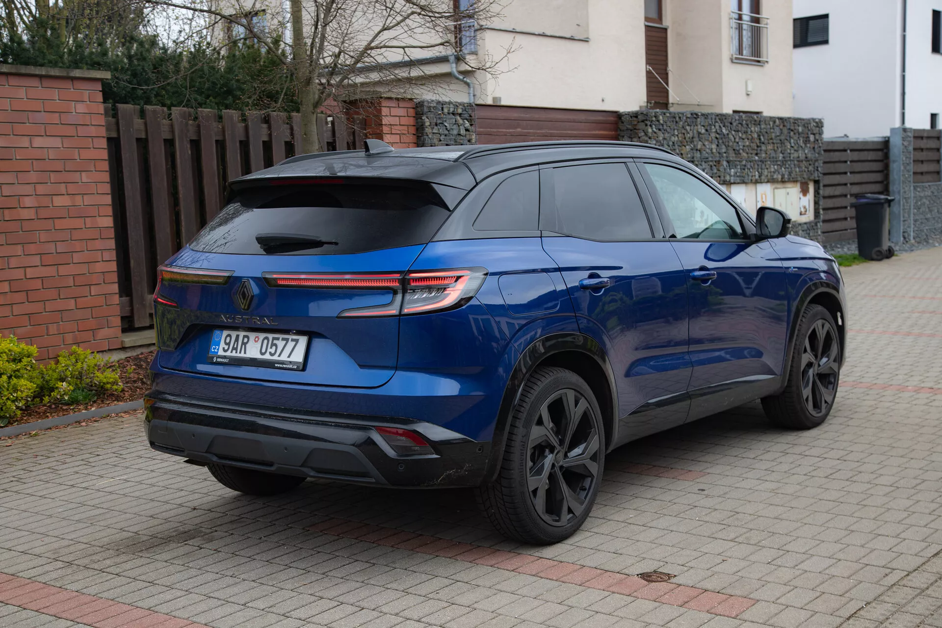 SUV moderno Renault Austral 2022 em azul metálico brilhante estacionado em uma entrada pavimentada ao lado de um edifício residencial, exibindo seu spoiler traseiro esportivo, lanternas traseiras LED, rodas de liga escura, janelas traseiras com privacidade e sensores de estacionamento de um ângulo traseiro de três quartos