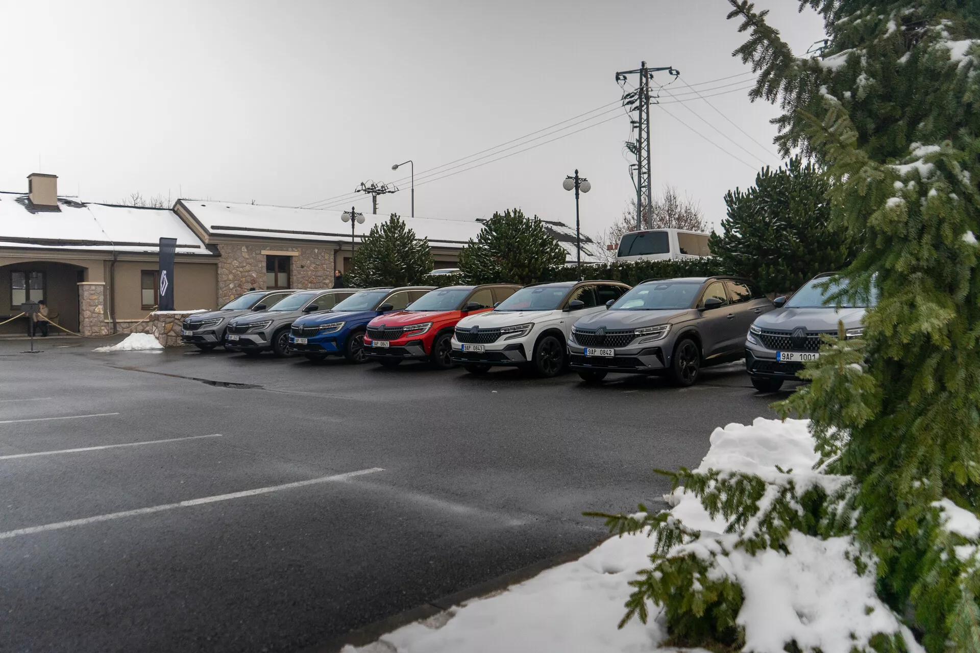 Linha de carros estacionados, incluindo sedãs e SUVs, sob uma leve queda de neve, indicando clima frio, com perfis laterais visíveis incluindo portas, espelhos laterais e rodas, tirada de um ângulo elevado da calçada