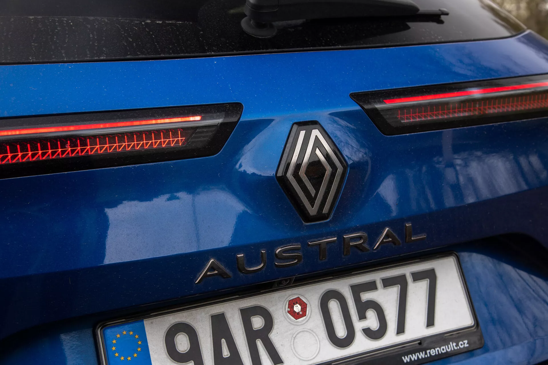 Imagem aproximada da secção traseira de um Renault Austral 2022 em azul brilhante, apresentando um logótipo em forma de diamante e o nome do modelo 'Austral' em letras maiúsculas, matrícula com a bandeira da União Europeia, distintivos faróis traseiros LED horizontais e possível sistema de iluminação traseira adicional, com o exterior do carro a indicar uso recente.