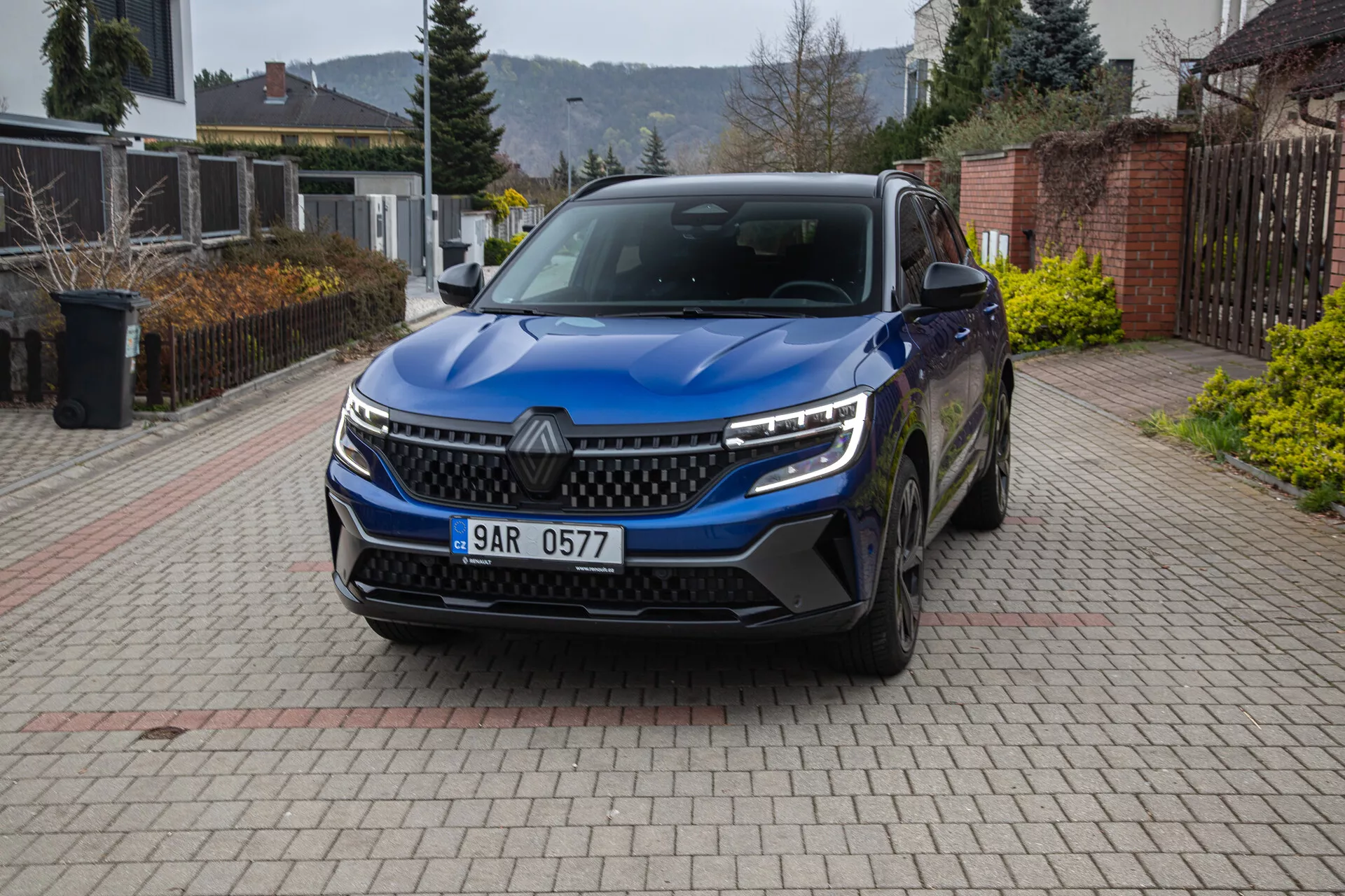 Vista frontal e angular de um SUV Renault Austral 2022 estacionado numa entrada de residência, mostrando a grade, faróis LED, luzes diurnas, para-choque com entradas de ar, sensores de estacionamento, emblema, rodas de liga, espelhos laterais com sinais de direção, e placa de número.
