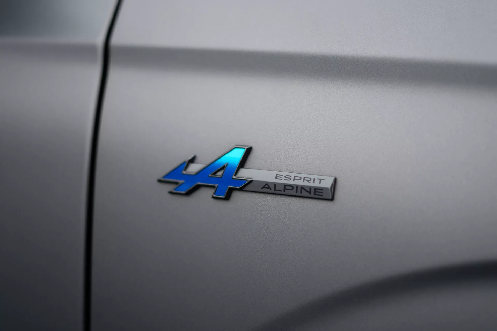 Imagem de close-up do emblema Alpine do Renault Austral 2022 com designação do modelo 'ALPINE ESPRIT' em superfície metálica azul e cromo, angulada para profundidade de campo reduzida