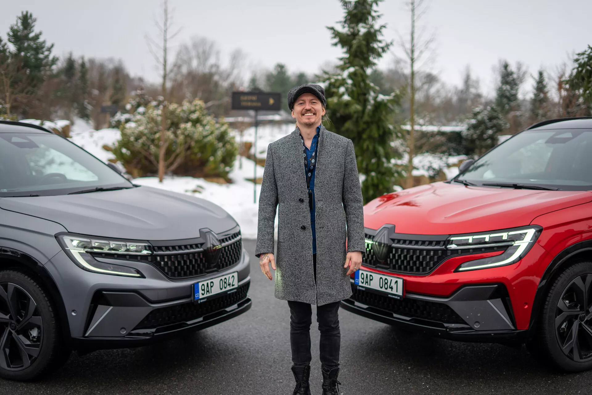 Pessoa sorridente ladeada por dois SUVs Renault Austral 2022 num cenário de inverno, destacando o design moderno dos veículos e as características tecnológicas avançadas com foco na grade frontal, faróis LED e perfil lateral elegante.