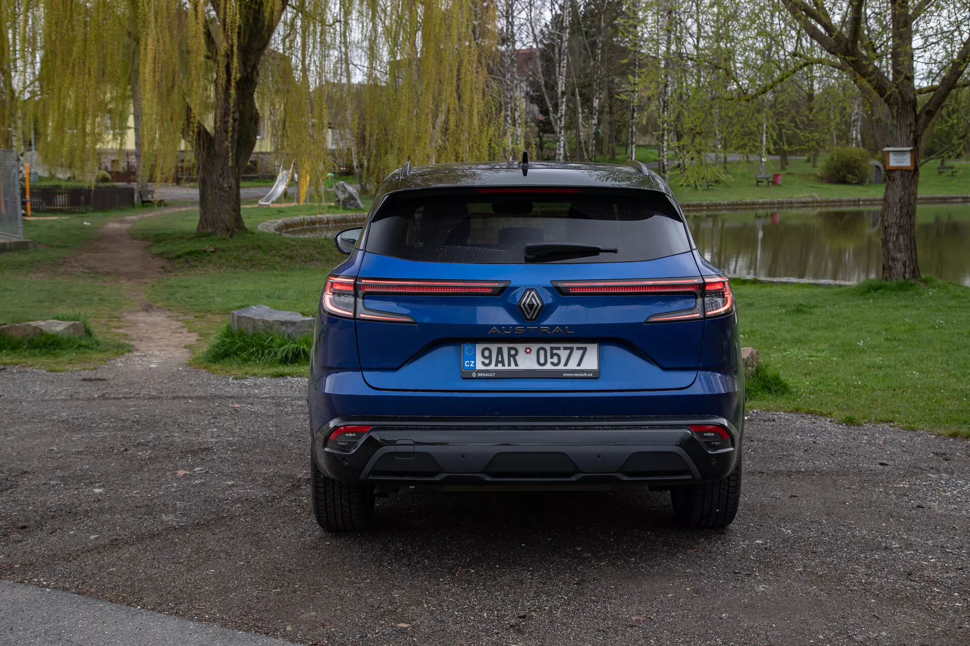 Vista traseira de um Renault Austral 2022 azul, estacionado ao ar livre, mostrando características incluindo o logotipo da marca, placa de licença, luzes traseiras modernas, para-brisa traseiro com limpador, para-choque traseiro projetado com possíveis sensores de assistência ao estacionamento, dicas de escape visíveis, refletores ou sensores, e um aerofólio traseiro para melhorar a aerodinâmica, tudo definido contra um fundo sereno semelhante a um parque com árvores, grama e um corpo de água.