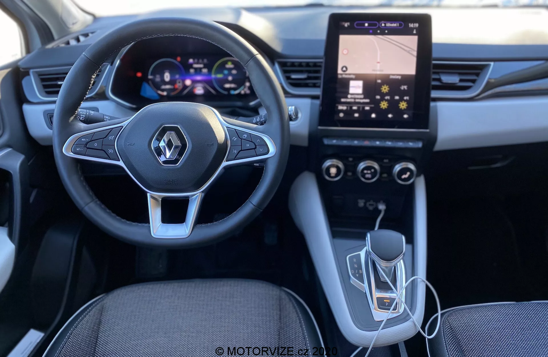 Imagem do interior do Renault Captur 2020 a partir da perspectiva do motorista, destacando o volante de design moderno com controles multimídia integrados, painel de instrumentos digital, sistema de entretenimento com tela sensível ao toque vertical e controles HVAC, alavanca de câmbio automática e conectividade USB. A iluminação e a qualidade sugerem um interior bem projetado e tecnologicamente avançado.