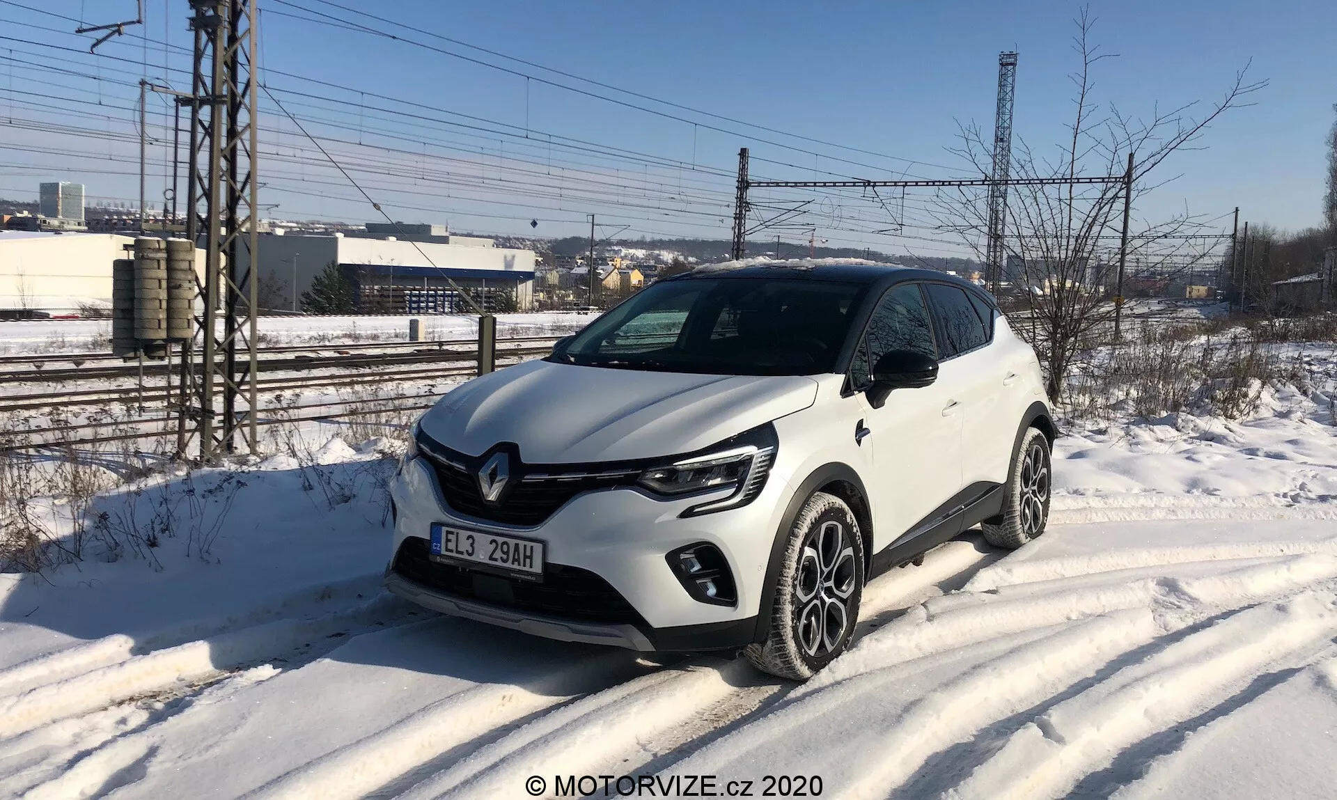 A imagem apresenta um Renault Captur 2020 branco, um SUV compacto estacionado à beira de uma estrada coberta de neve, capturado de um ângulo frontal de três quartos. O veículo exibe luzes diurnas de LED em forma de C, faróis projetores, uma grade dianteira com acentos pretos e um distintivo proeminente da Renault, e rodas de liga leve multi-raios. Elementos de design notáveis incluem revestimento de plástico ao longo das bordas inferiores e dos arcos das rodas. O ambiente sugere um dia frio de inverno com um fundo industrial ou semiurbano.
