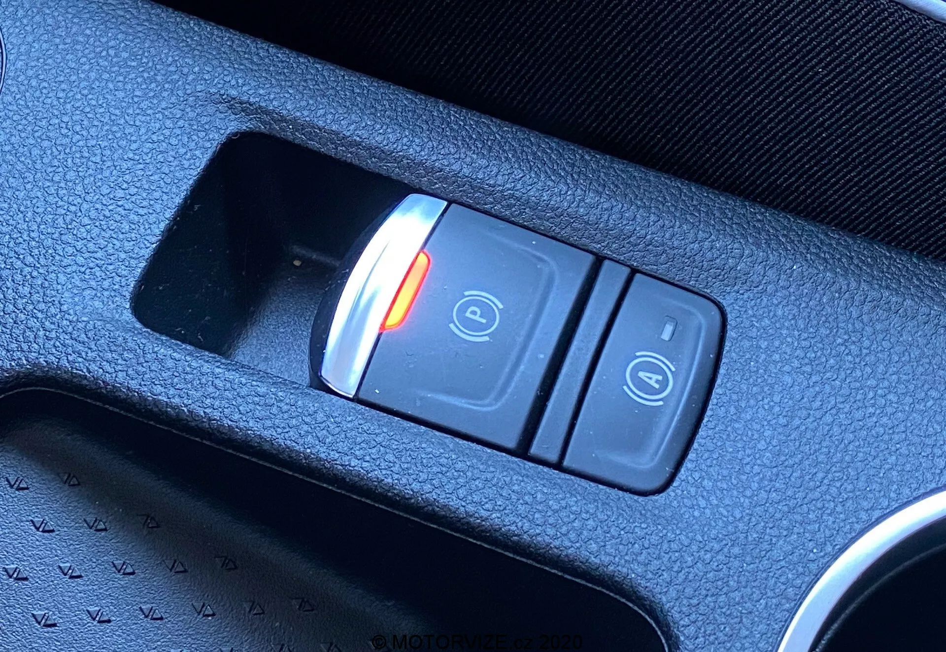 Close-up diagonal do interruptor do travão de estacionamento eletrónico no interior do Renault Captur 2020. O interruptor, localizado na consola central, apresenta dois botões: um botão vermelho iluminado com um cadeado fechado e o símbolo 'P' indicando o estado ativado, e outro botão com o símbolo do cadeado aberto. O interruptor está inserido numa consola de plástico texturizado preto, destacando a tecnologia moderna do veículo.