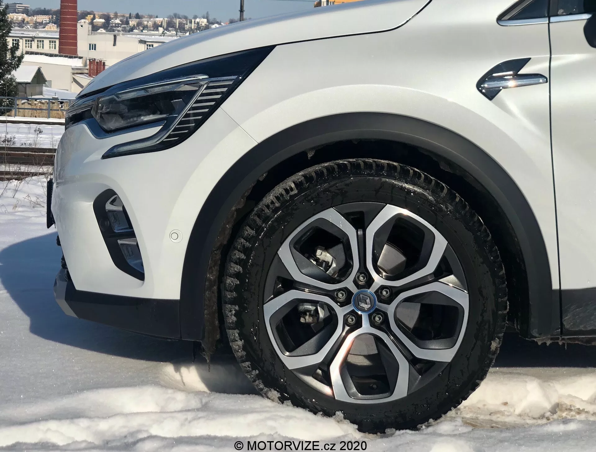 Close-up de um Renault Captur 2020 a partir de um ângulo baixo mostrando a seção dianteira esquerda, com foco na roda de liga, para-lama, para-choque e área do farol. O carro está sobre uma superfície nevada em um ambiente urbano com prédios e céu claro.