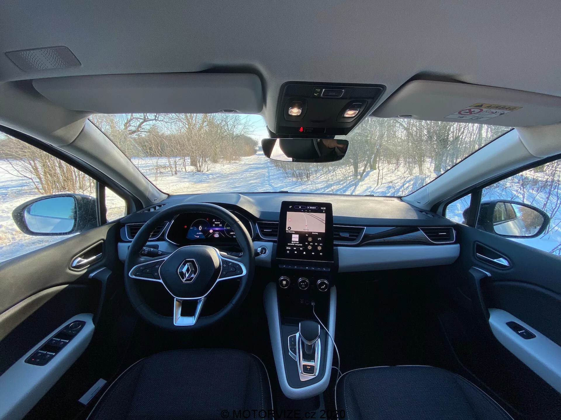 Imagem do interior do Renault Captur 2020 a partir dos bancos traseiros mostrando a cabine frontal com volante de três raios com botões integrados, painel de instrumentos digital, sistema de infotainment com ecrã tátil verticalmente orientado, consola central moderna com poucos controlos físicos, seletor de mudanças de design elegante, saídas de ventilação de HVAC, materiais de alta qualidade incluindo couro e acentos metálicos, espelho retrovisor possivelmente com sensores para ADAS e pilar A visível com controlos de janelas.