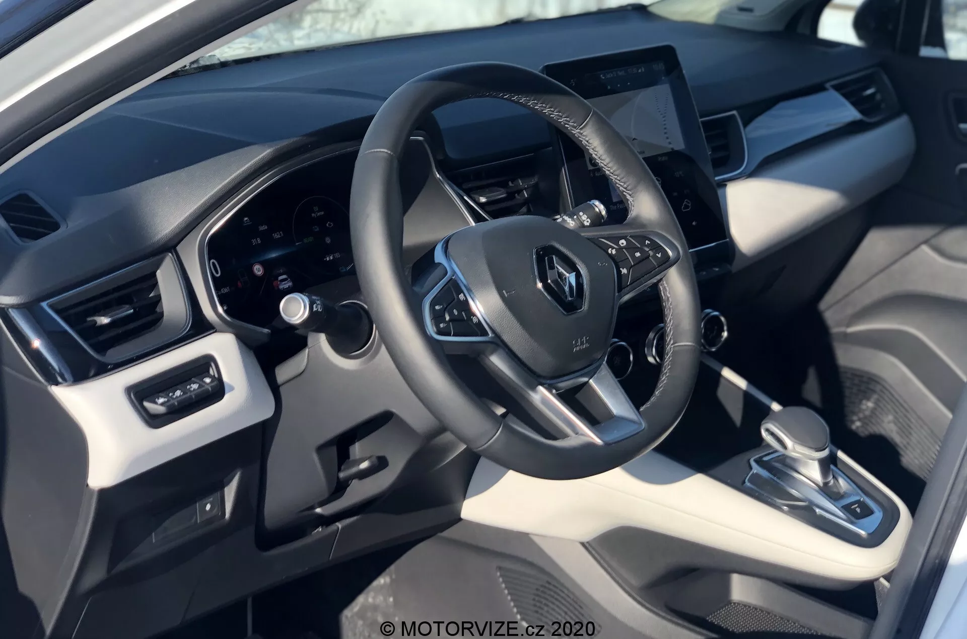 Vista interior do Renault Captur 2020 da perspectiva do motorista, mostrando o volante com o logotipo da Renault, controles montados, painel de instrumentos digital atrás e um sistema de infoentretenimento com tela sensível ao toque centralmente localizado. A imagem destaca uma mistura de materiais de couro sintético e plástico com acentos metálicos, transmissão automática, sistema de controle climático e ajustes dos vidros e espelhos elétricos.