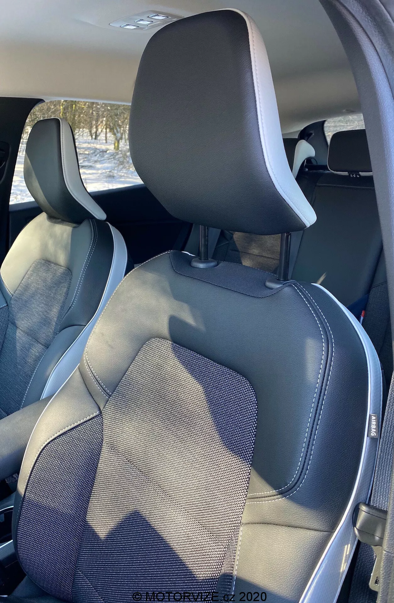 Vista do interior do Renault Captur 2020, mostrando os assentos do passageiro dianteiro e do motorista a partir de um ângulo traseiro, ligeiramente deslocado para a direita. A seção superior dos assentos inclui encostos de cabeça integrados e molduras visíveis dos cintos de segurança. Os assentos apresentam um design moderno de duas tonalidades com cores cinza escuro e claro e bordas em couro sintético com costuras detalhadas, destacando artesanato e estilo.