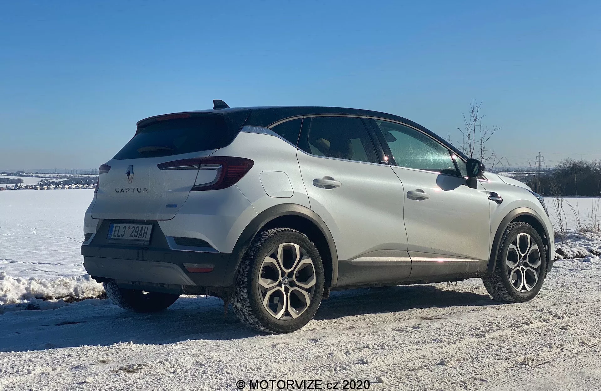 Imagem de um Renault Captur 2020, um SUV compacto, em um ambiente de inverno. Exibido de uma vista traseira de três quartos, o carro apresenta um design elegante com linhas suaves e luzes traseiras de LED. O emblema do fabricante e o nome do modelo são visíveis juntamente com rodas de liga leve de múltiplos raios, sensores de estacionamento traseiros e espelhos laterais com setas integradas. Posicionado em uma área de cascalho coberta de neve, a imagem sugere habilidade em vários terrenos e condições de inverno.