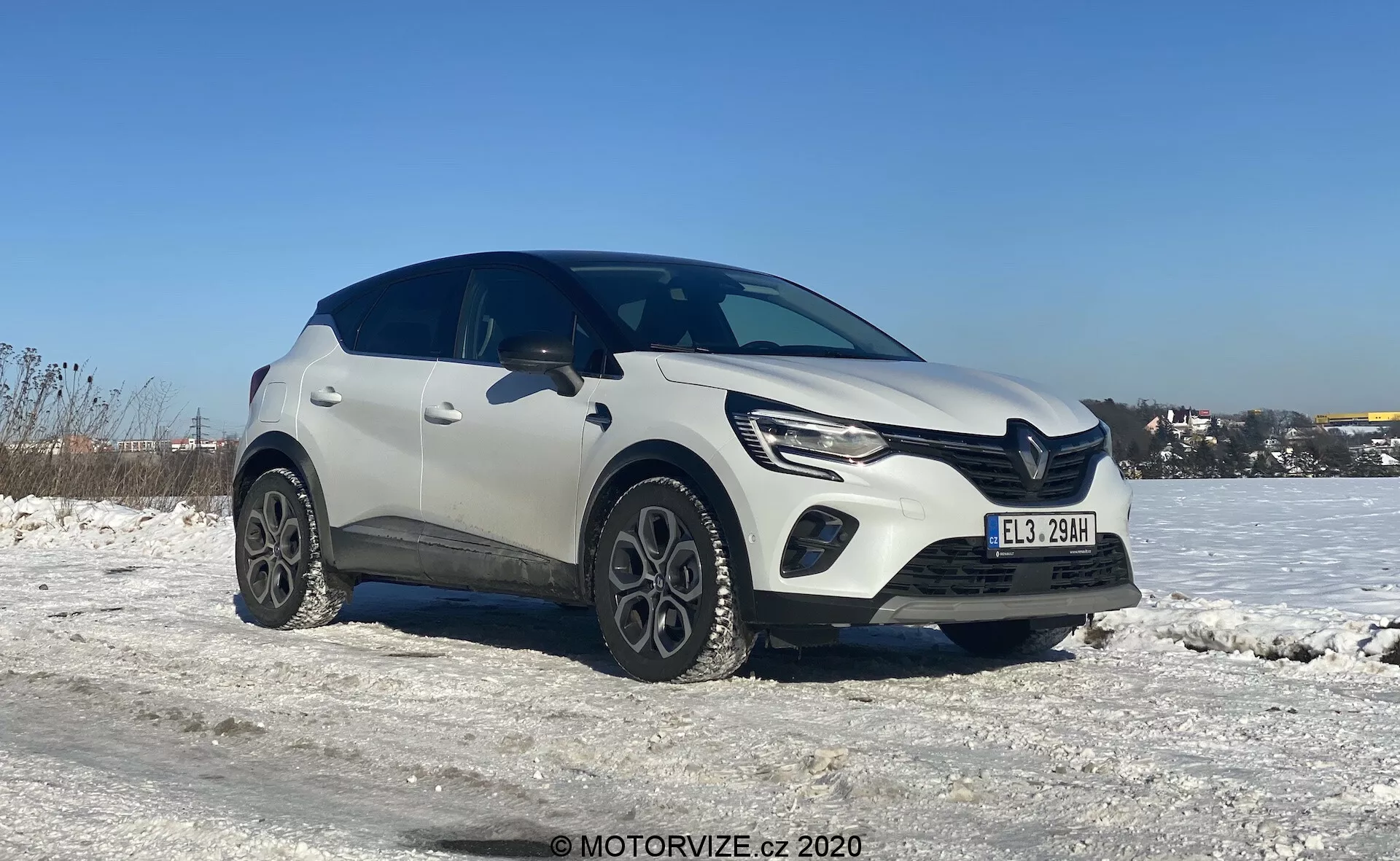 Vista em três quartos de um SUV crossover compacto Renault Captur 2020 branco em um ambiente de inverno, com chão coberto de neve e céu azul claro. A imagem destaca o lado dianteiro esquerdo e parte do perfil lateral do veículo, tirada de um ângulo ligeiramente inferior que enfatiza as rodas e a metade inferior. O design moderno do carro mostra linhas distintas no capô, faróis LED elegantes com luzes diurnas e uma grade dianteira proeminente. Rodas de liga únicas e revestimento plástico ao redor dos arcos das rodas adicionam um visual robusto. As sombras sob o carro sugerem que a foto foi tirada ao meio-dia ou no início da tarde com luz natural do dia.