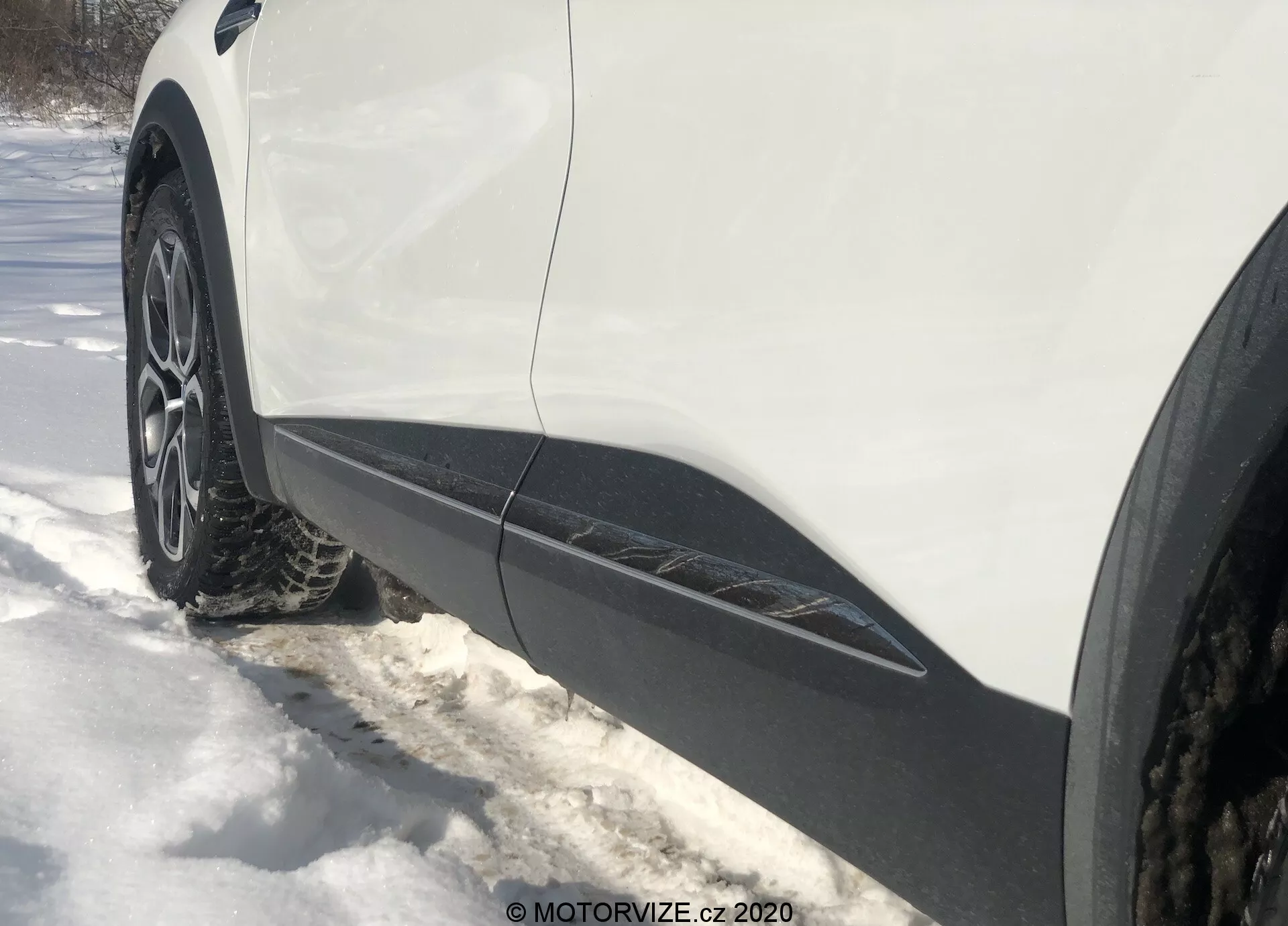 Vista lateral de perto, angulada a partir do nível do solo da roda dianteira e para-lamas do lado do motorista de um Renault Captur 2020. A roda multi-raios de liga com pneu de inverno ou para todas as estações está cercada de neve. A carroceria branca com acabamento preto corre ao longo da borda inferior, indicando um crossover ou SUV. A iluminação sugere um dia ensolarado com sombras visíveis.