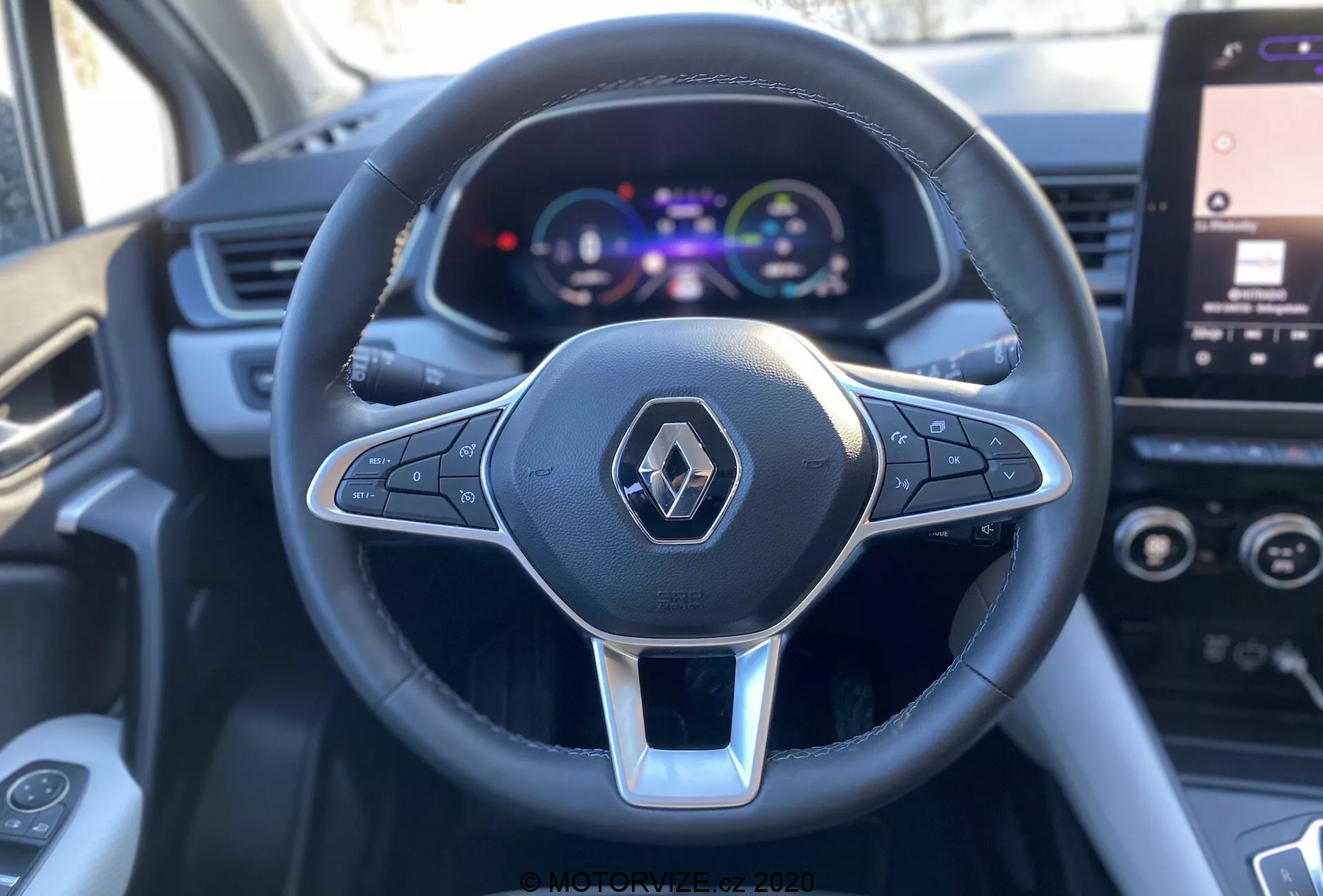 Close-up, visão em primeira pessoa do banco do motorista de um Renault Captur 2020, focando no volante multifuncional de três raios revestido em couro com detalhe prateado e logotipo da Renault, juntamente com um painel de instrumentos digital colorido exibindo velocímetro e detalhes do veículo, com uma tela de infotainment parcialmente visível ao fundo.