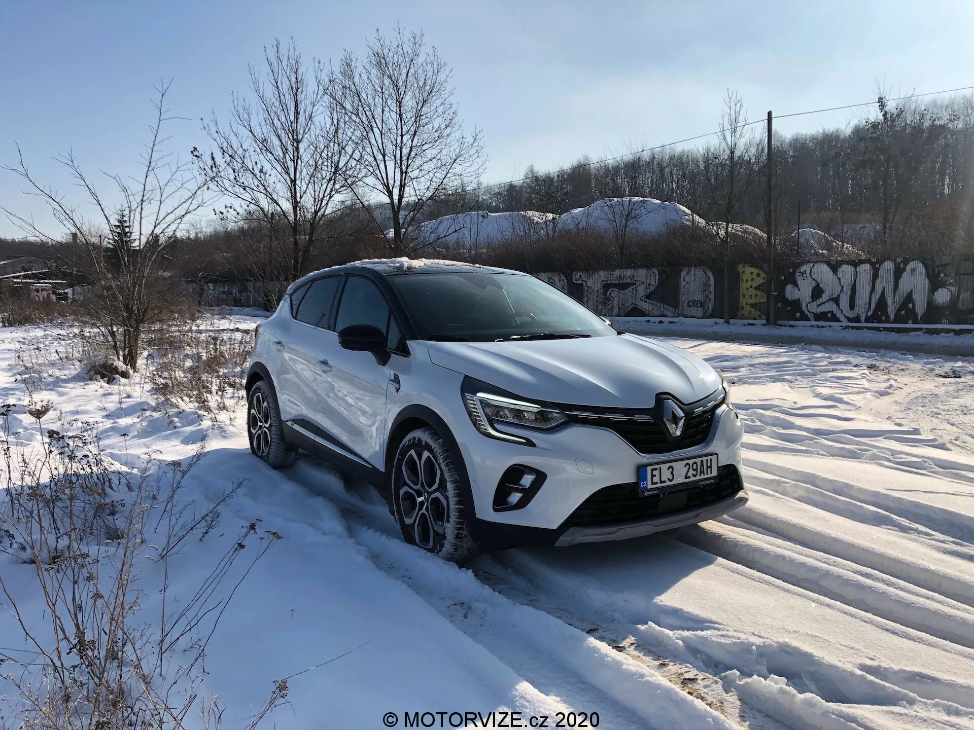 Um crossover SUV Renault Captur 2020 branco está estacionado sobre um solo coberto de neve com vegetação e estruturas cobertas de grafite ao fundo, sob um céu predominantemente claro com algumas nuvens. A imagem é tirada de um ângulo de três quartos focado no lado do motorista, mostrando a grade frontal com um emblema cromado e faróis de LED contemporâneos. O SUV apresenta luzes diurnas LED frontais, alojamentos de luz de neblina, linhas de caráter no capô, revestimento plástico ao redor dos arcos das rodas, espelhos laterais com sinalizadores de direção integrados, rodas de liga com múltiplos raios e pneus de inverno, e uma linha de ombro forte. A postura elevada implica maior distância ao solo, adequada para diversos terrenos.