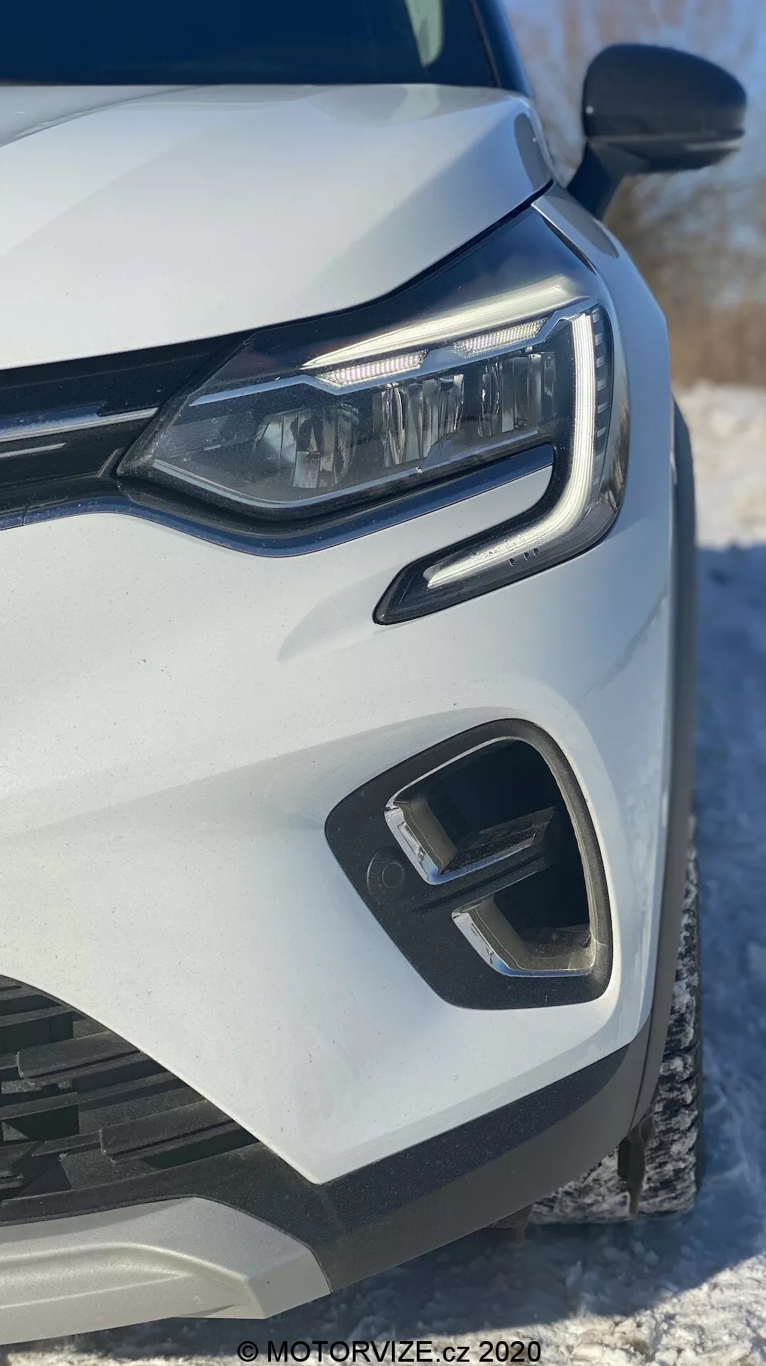 Close-up da parte dianteira esquerda de um Renault Captur 2020 branco, focando no farol LED moderno, para-choque dianteiro geométrico e elementos da grade, capturado de um ângulo frontal baixo com neve ao fundo