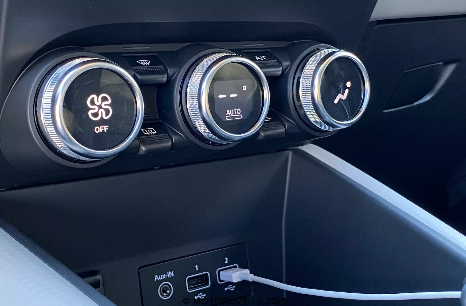 Close-up do interior do Renault Captur 2020 focado no sistema de controle climático. A imagem mostra três grandes discadores no painel de instrumentos. O primeiro discador controla o ar-condicionado, indicado por um símbolo de floco de neve e um botão 'OFF'. O discador do meio regula a temperatura com gradientes de cor vermelho e azul e uma indicação 'AUTO'. O terceiro discador com um ícone de ventilador ajusta a velocidade do ventilador. Abaixo dos discadores, há duas portas USB e uma entrada auxiliar, com um cabo conectado a uma das portas USB. Os materiais são de plástico ou sintéticos escuros com acabamento fosco e possuem um design moderno e minimalista.