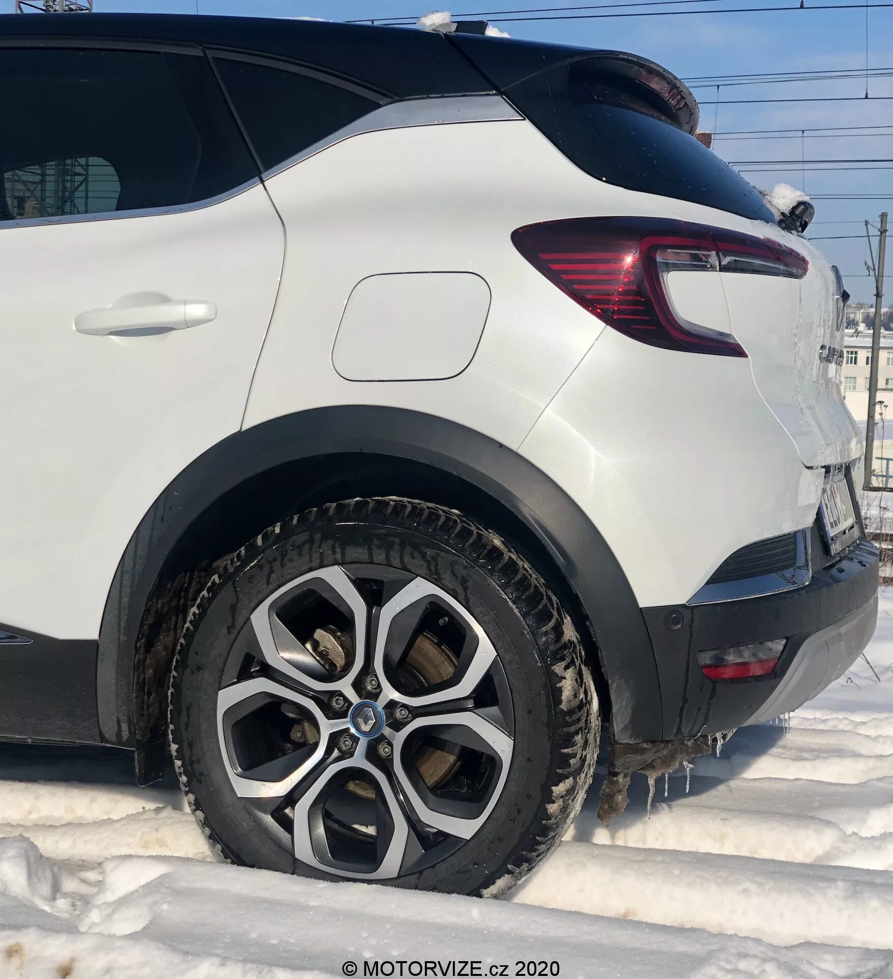Close-up detalhado de um Renault Captur 2020 branco, mostrando o lado do passageiro traseiro de um ângulo baixo. Destaques incluem a roda traseira com design de múltiplos raios de liga, suja de lama e neve, sugerindo condições de inverno. A imagem captura o para-lama traseiro limpo acima do arco da roda, a moderna lanterna traseira com seções de LED vermelhas e claras, e a proteção robusta de plástico atrás da roda. A janela inclinada na porta traseira contribui para a forma aerodinâmica do veículo.