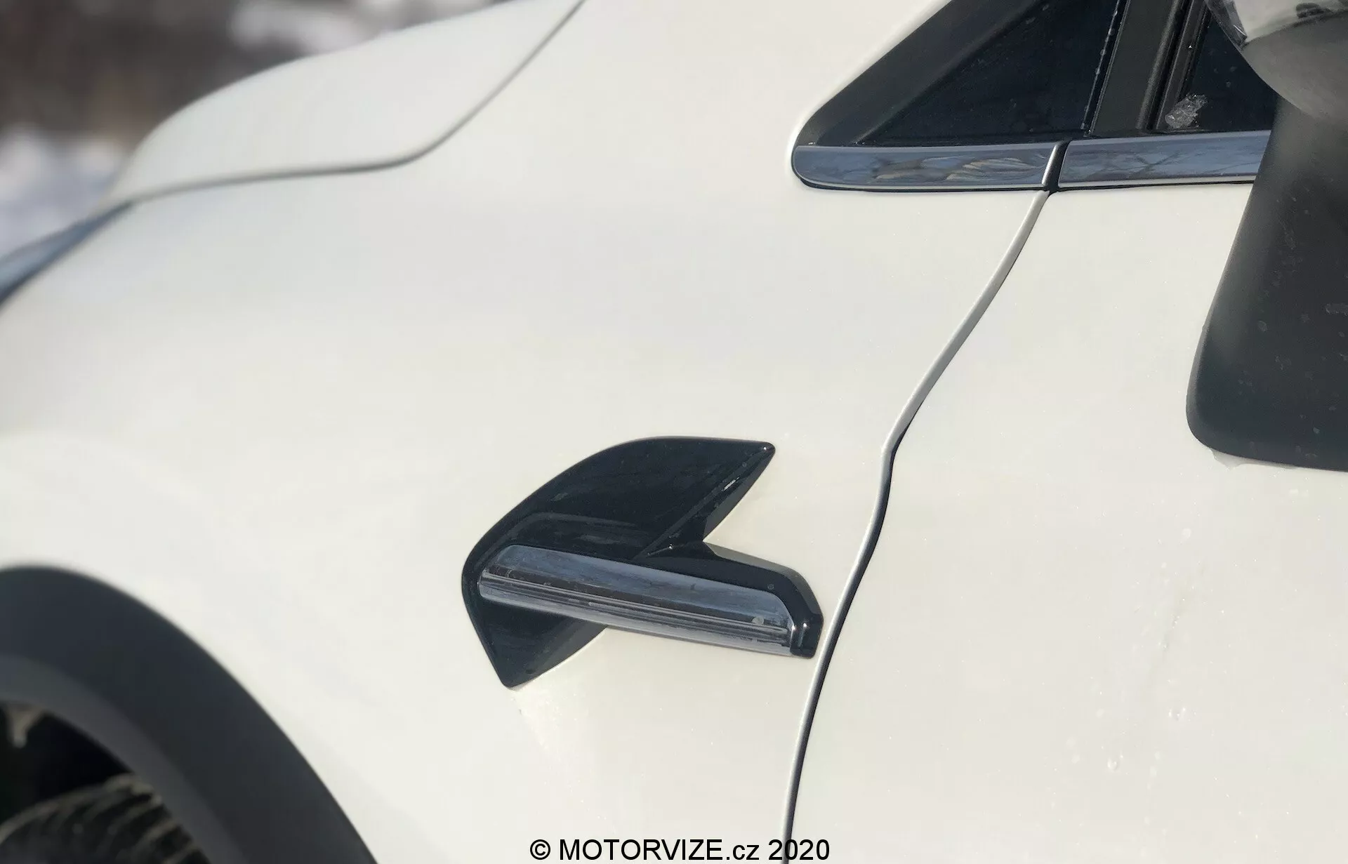 Close-up do exterior do Renault Captur 2020, focando no lado do veículo branco. A imagem apresenta uma antena preta em forma de barbatana de tubarão no topo esquerdo, projetada para recepção de rádio e aerodinâmica. Centralizado abaixo da antena, um duto ou entrada de ar preta é destacado, possivelmente para resfriamento dos freios ou melhoria do fluxo de ar. A linha aerodinâmica do teto do carro curva-se para baixo em direção à traseira, com a pintura branca brilhante refletindo a luz. O fundo está levemente desfocado, indicando um cenário externo com neve.