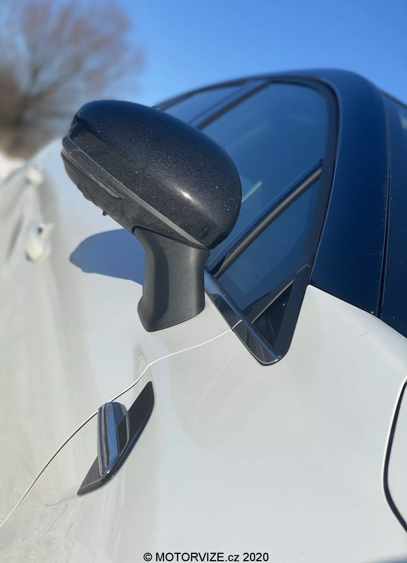 Vista aproximada da montagem do espelho lateral do Renault Captur 2020 da frente para trás, focando na carcaça de plástico texturizado preto com sinal de pisca integrado e refletindo o céu azul. O painel elegante de cor escura abaixo e o possível espelho de ponto cego indicam um design moderno e recursos avançados.