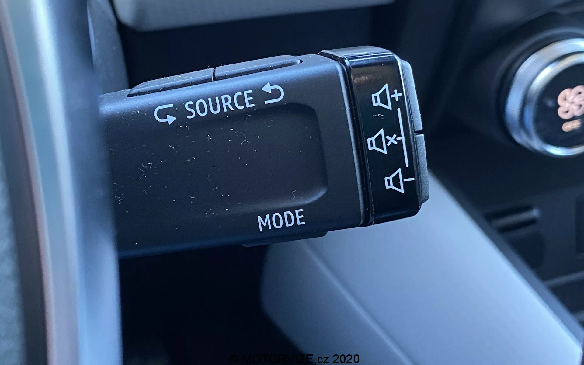 Close-up dos controles de áudio montados no volante de um Renault Captur 2020, focando na alavanca de controle do lado esquerdo com botões para SOURCE, ajuste de volume, pular faixas e seleção de MODE. A fotografia captura os detalhes dos botões de plástico com acabamento fosco e seu design ergonômico, com parte da coluna de direção e interior do carro desfocados ao fundo, destacando a funcionalidade e conveniência para o motorista.