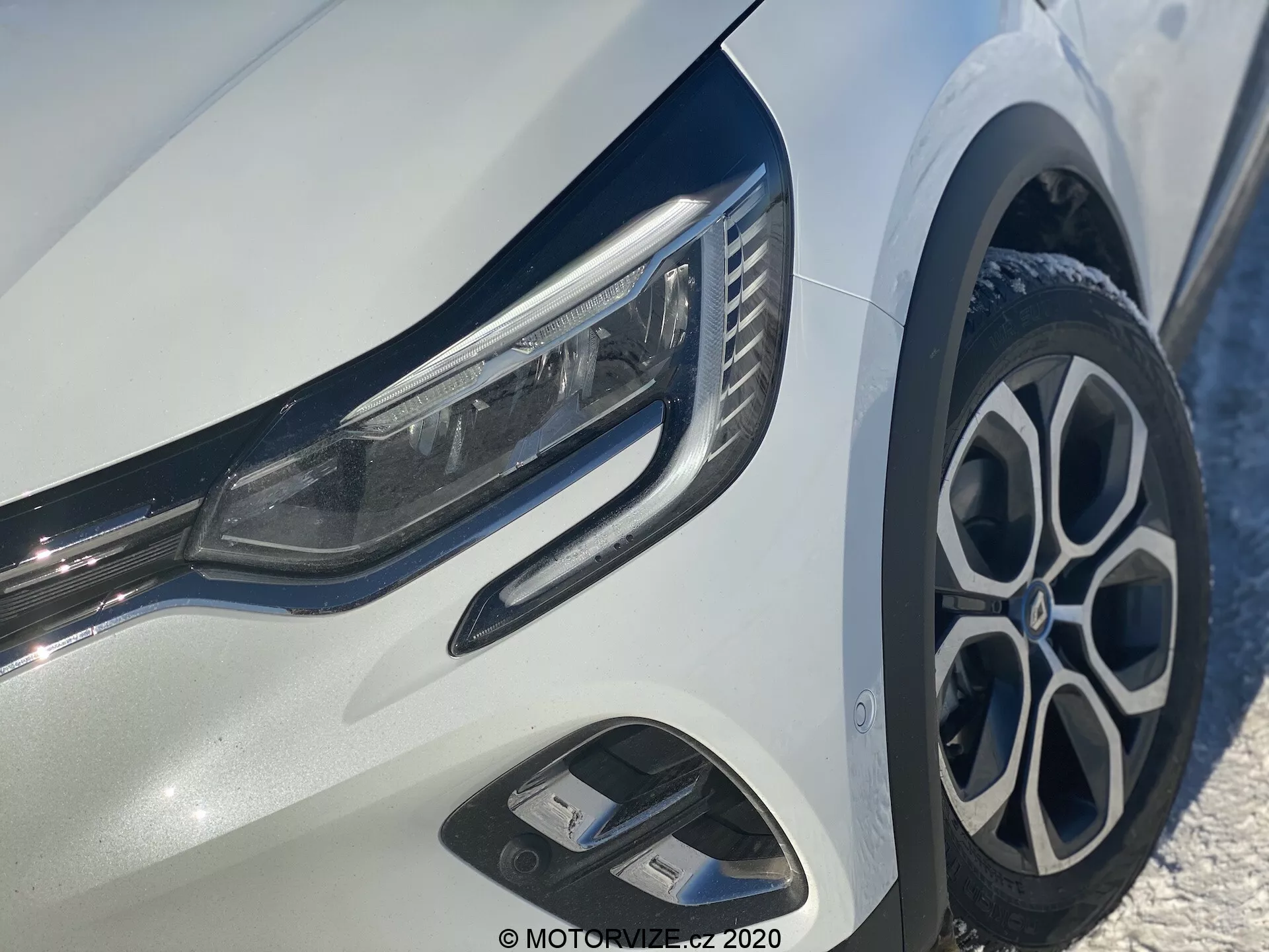 Vista em close-up do Renault Captur 2020 focalizando o quarto frontal esquerdo, mostrando faróis LED elegantes, rodas de liga leve com vários raios, para-choque dianteiro dinâmico com entradas de ar e acabamentos cromados brilhantes em uma carroceria branca brilhante.